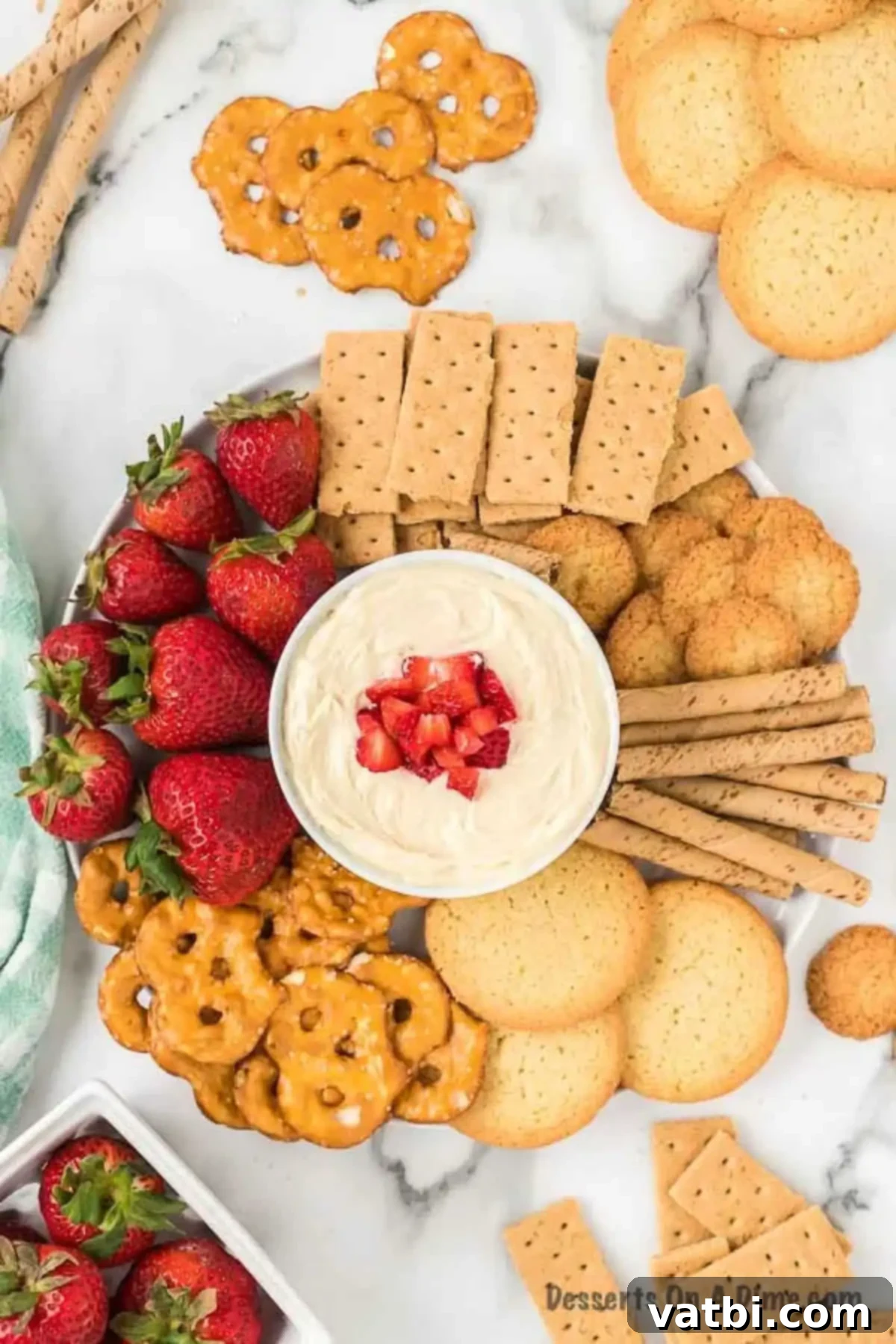 A festive serving tray showcasing a bowl of creamy cheesecake dip, accompanied by an array of dippers including graham crackers, various cookies, crispy pretzels, and fresh, vibrant strawberries.