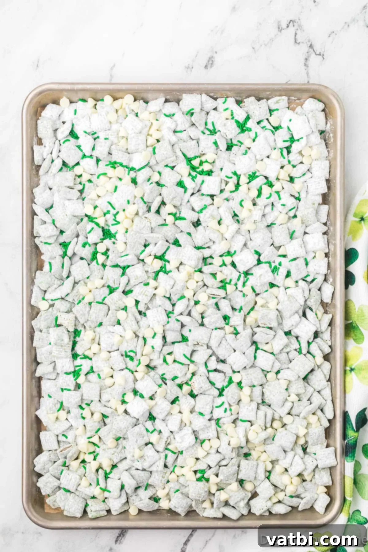 St. Patrick's Day Puppy Chow coated in powdered sugar, topped with white chocolate chips, green M&Ms, and green sprinkles, ready for cooling.