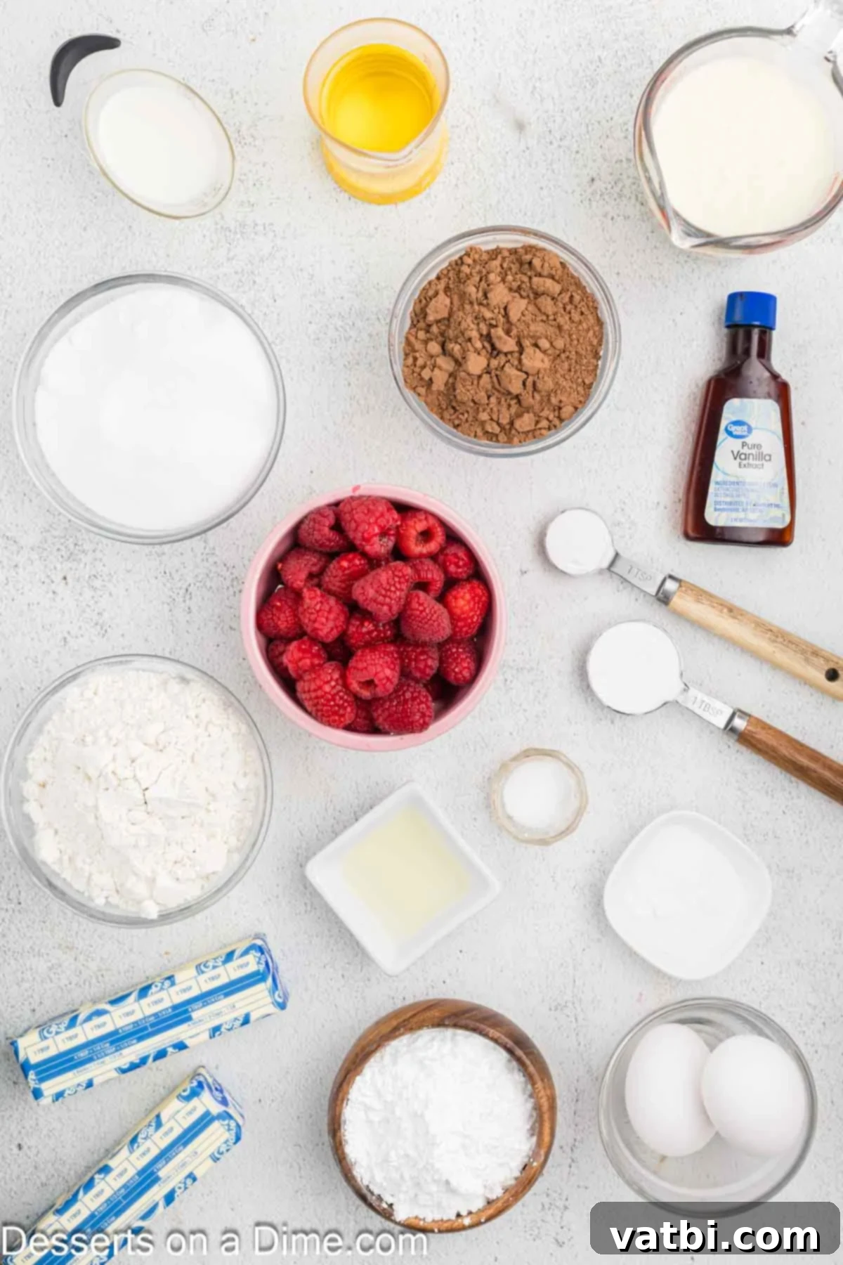 Decadent Chocolate Raspberry Cake 3 Various ingredients for chocolate raspberry cake including flour, cocoa powder, baking soda, buttermilk, eggs, vanilla, cornstarch, fresh raspberries, lemon juice, powdered sugar, and milk laid out on a surface.
