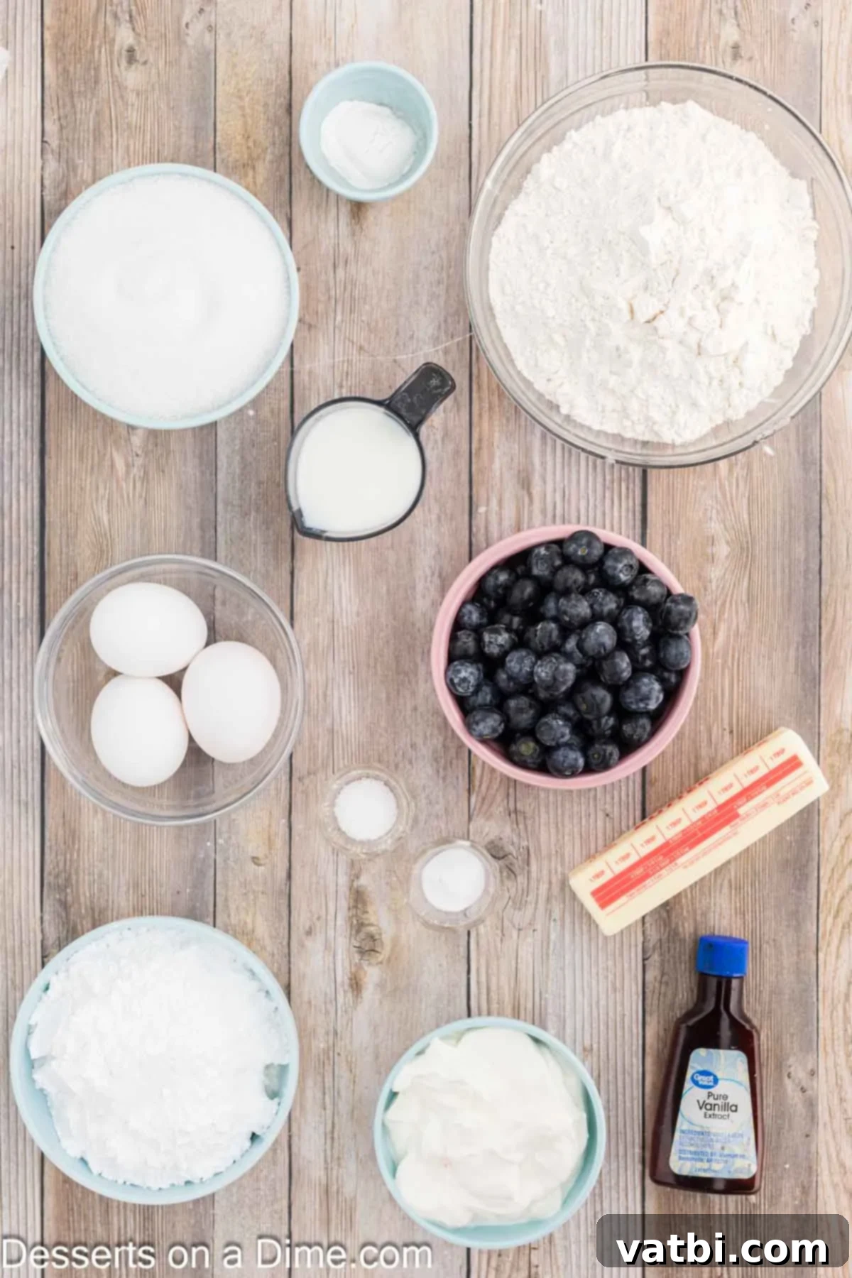 Blueberry Crown Cake 3 A selection of fresh ingredients laid out for baking a Blueberry Bundt Cake: a bowl of all-purpose flour, baking soda, softened unsalted butter, fresh large eggs, creamy full-fat sour cream, and plump blueberries.