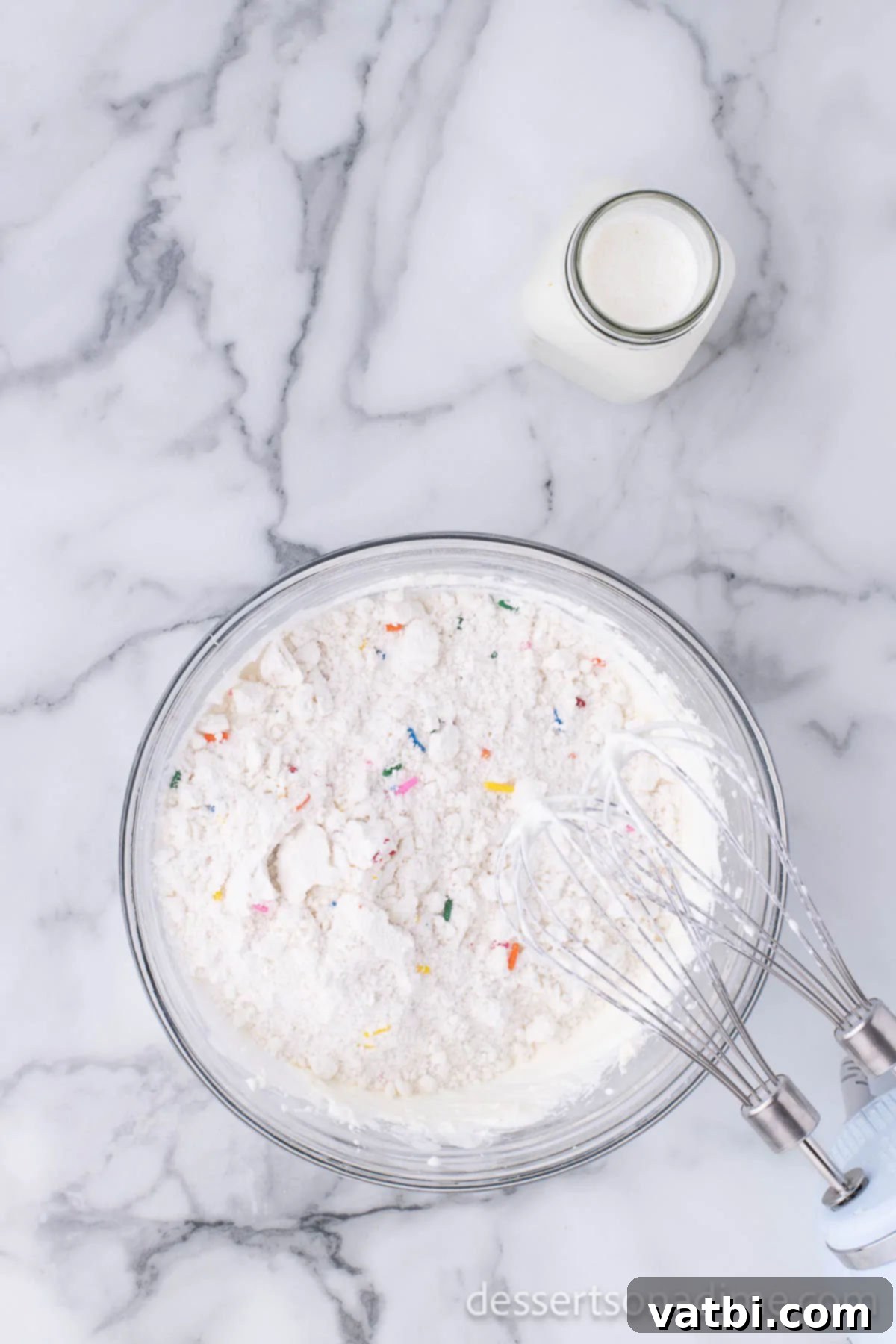 Party Cake Batter Delight 6 Funfetti cake mix being added to the blended cream cheese and Cool Whip mixture in a large mixing bowl, alongside half of the milk, prior to mixing.