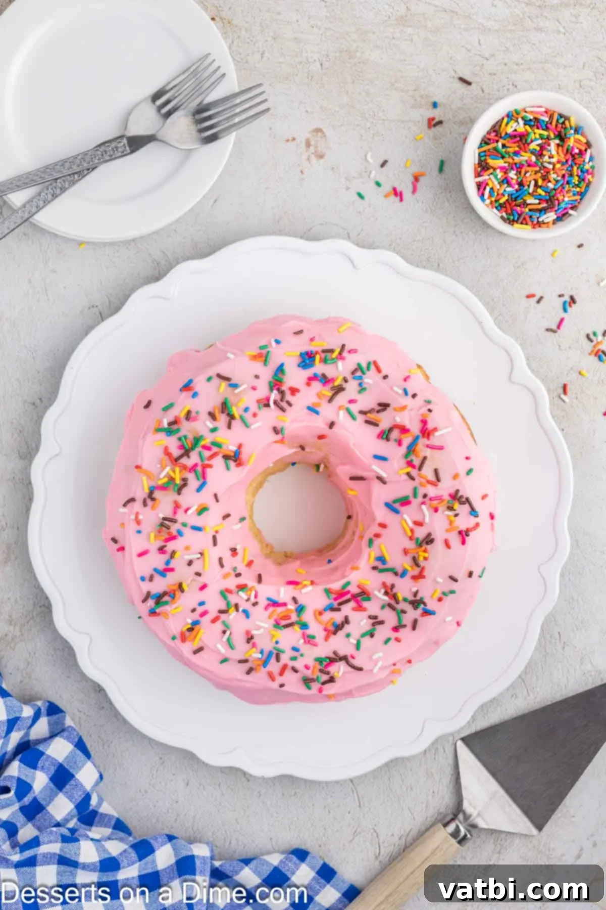 Donut Cake Delight 10 A whole Donut Cake, exquisitely glazed with pink icing and adorned with vibrant sprinkles, resting elegantly on a pristine white serving platter.