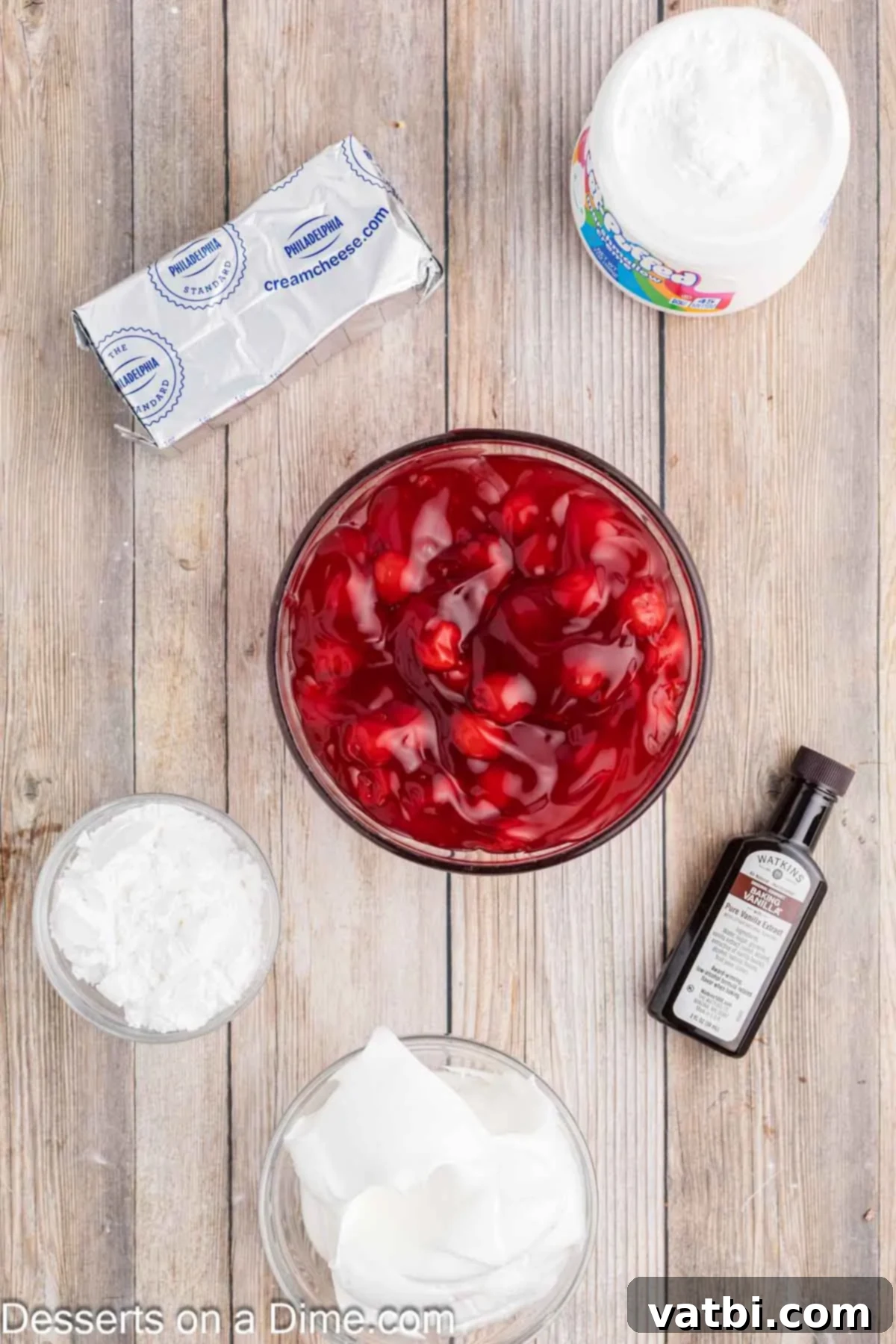 Creamy Cherry Dream Dip 3 Ingredients laid out for Cherry Cheesecake Dip: a block of cream cheese, powdered sugar, a bottle of vanilla extract, a jar of marshmallow creme, a tub of whipped topping, and a can of cherry pie filling.