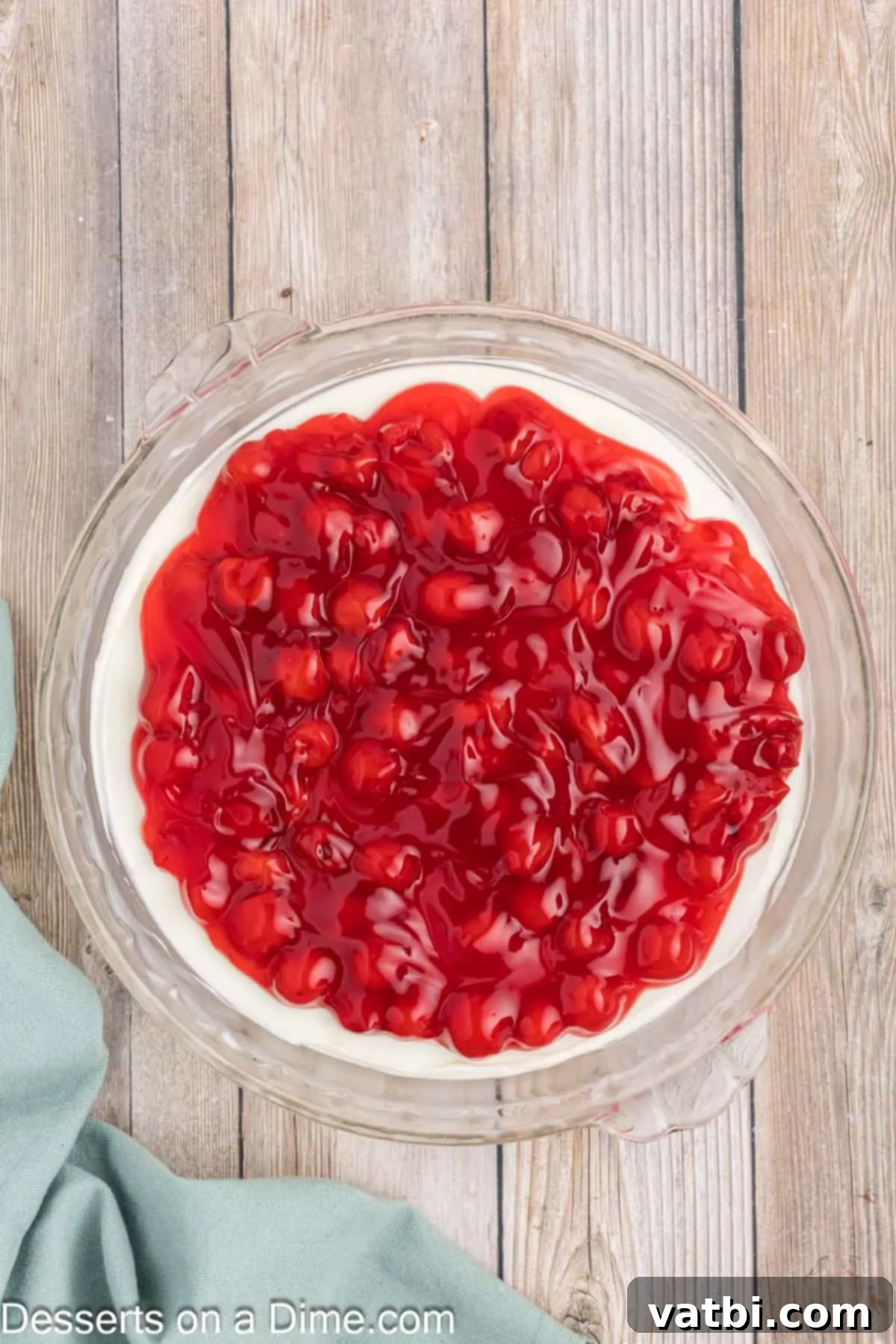 Creamy Cherry Dream Dip 7 Vibrant cherry pie filling being carefully spooned and spread evenly over the creamy white cheesecake base in a glass serving dish.