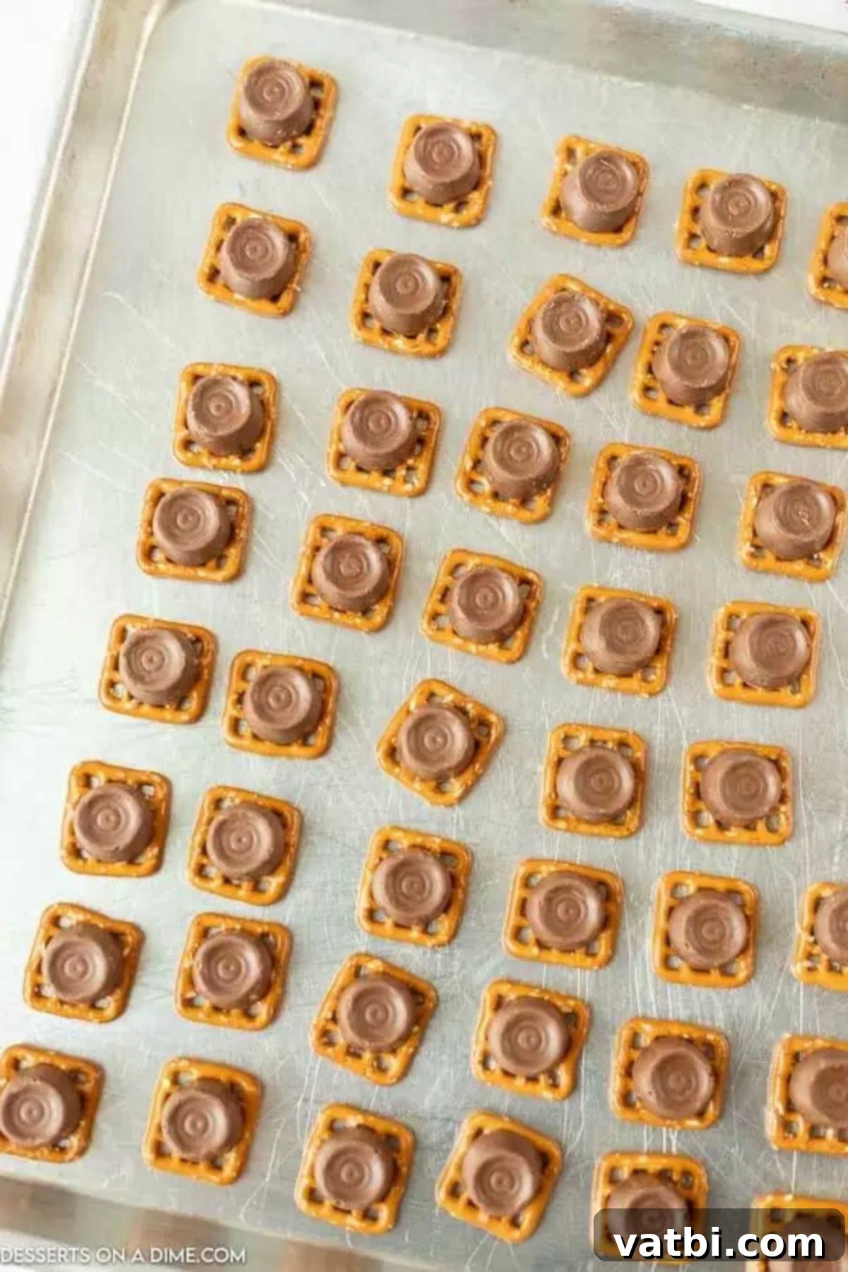 Square pretzels neatly arranged on a parchment-lined baking sheet, each topped with an unwrapped Rolo candy, ready for baking.