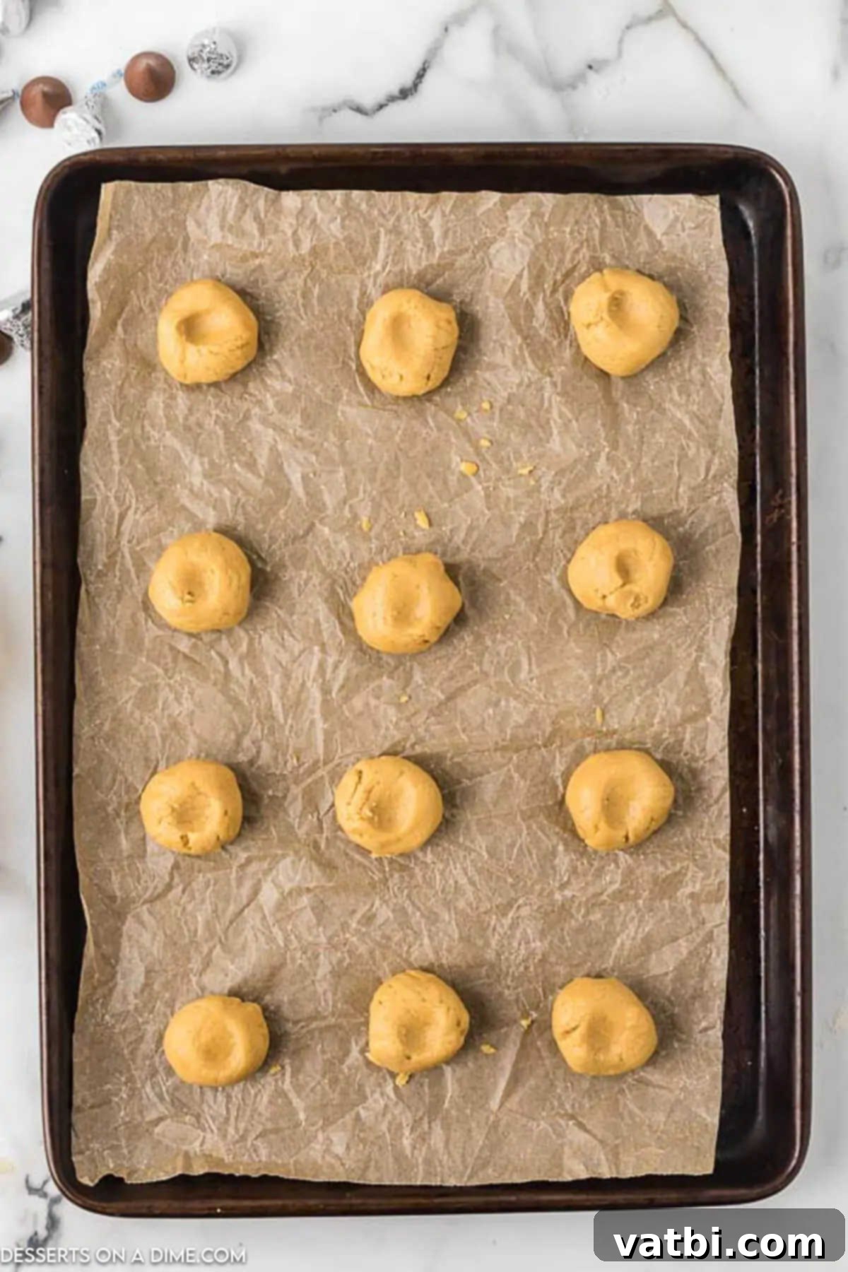 Chocolate-Kissed Peanut Butter Delights 7 Peanut butter dough balls with indention in the center on a baking sheet, ready for the oven.