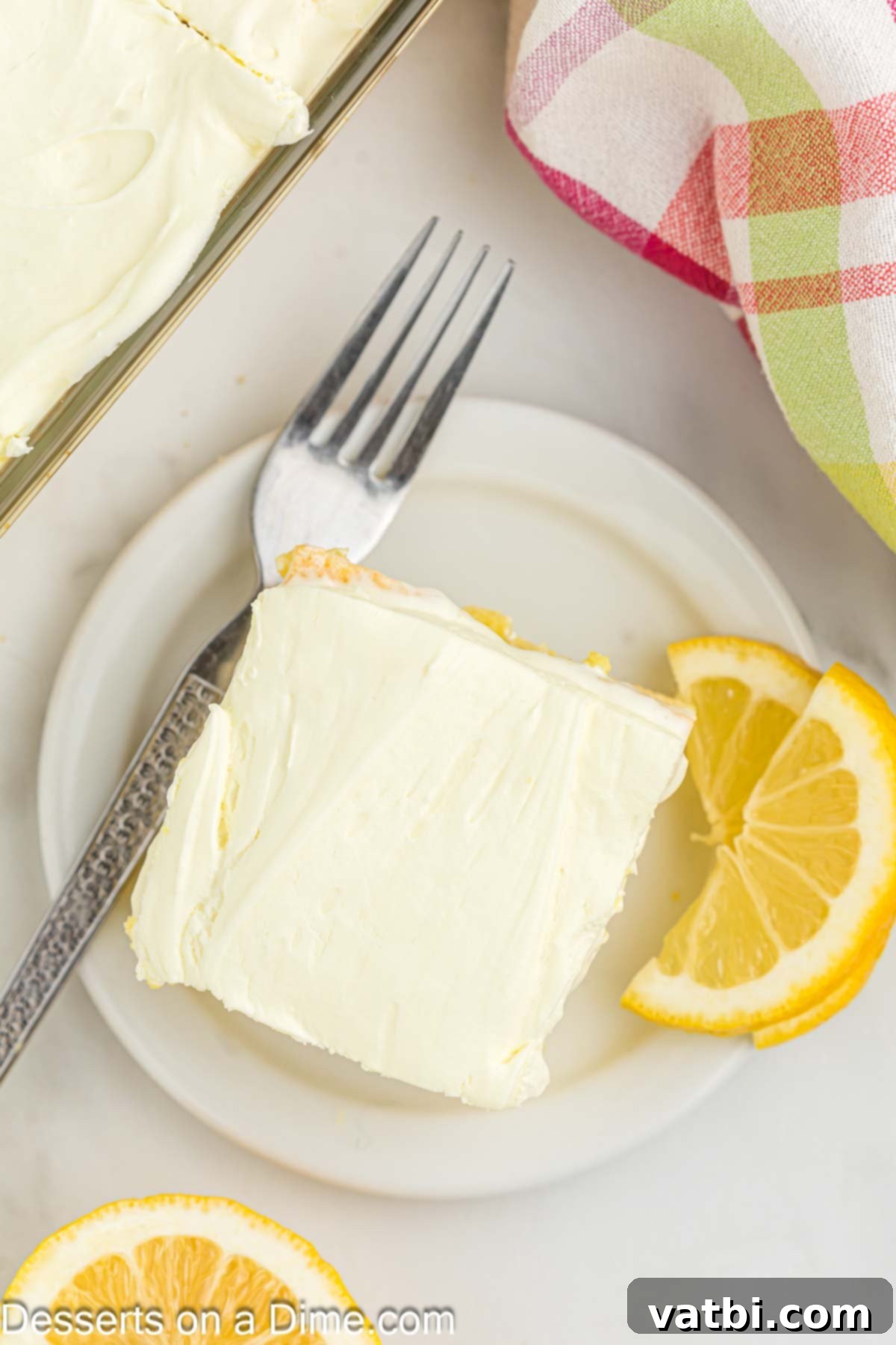 Tangy Lemon Infusion Cake 10 A generous slice of moist lemon poke cake on a white plate, showcasing the distinct layers of cake, Jell-O, and creamy frosting, garnished with a lemon slice.