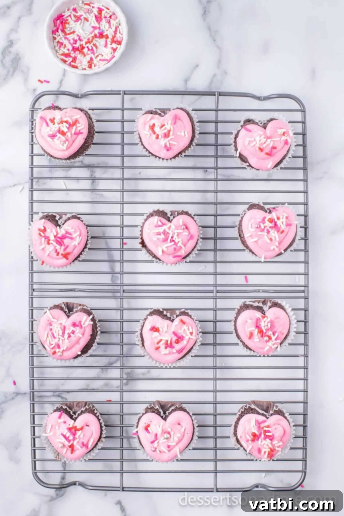 Red, pink, and white sprinkles being added to the pink-frosted heart-shaped cupcakes.