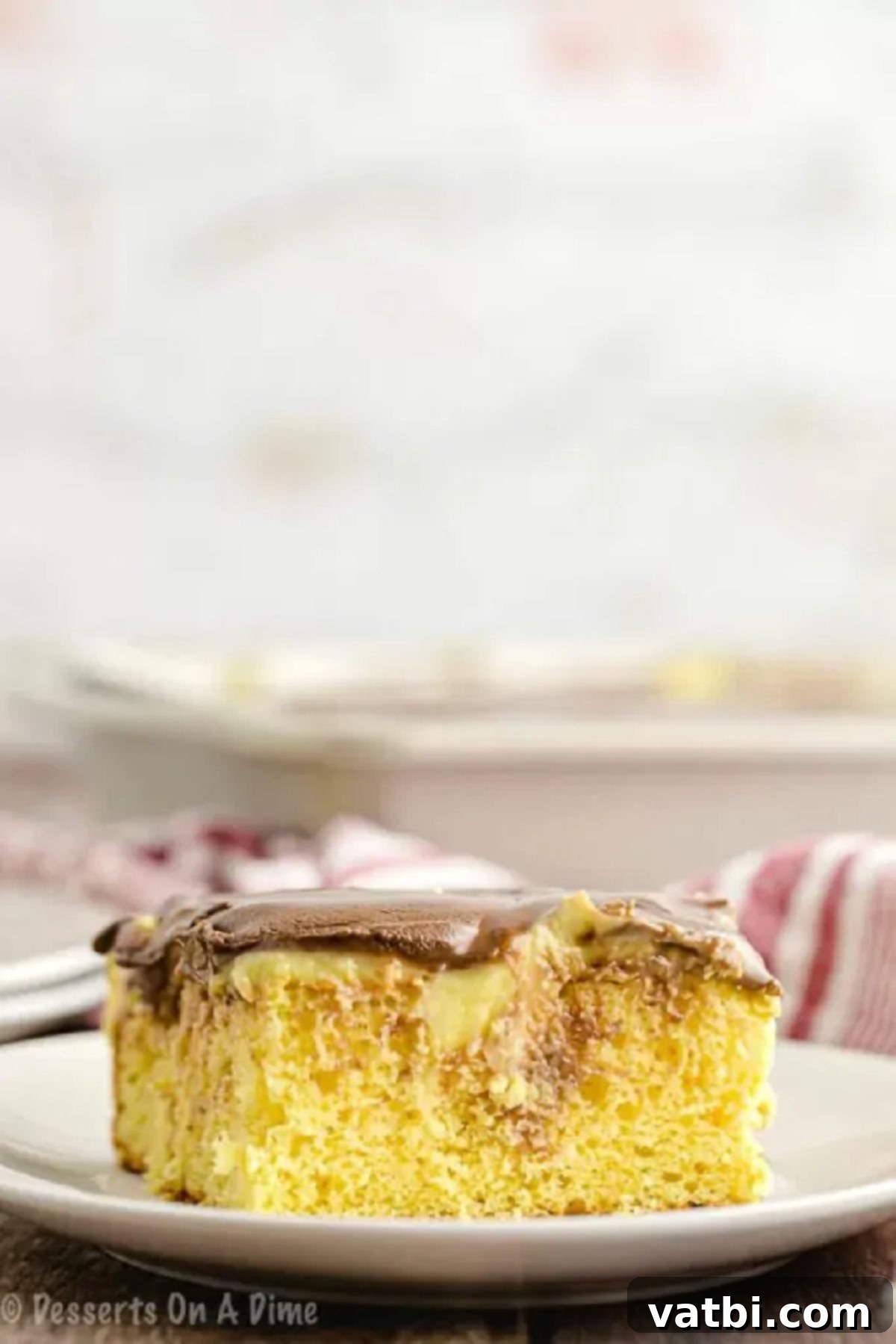 Boston Cream Poke Cake recipe is easy to make and full of flavor. Get ready for a delicious dessert that everyone will love! A slice of the Boston Cream Poke Cake on a white serving plate, showing the layers of cake, pudding, and chocolate.