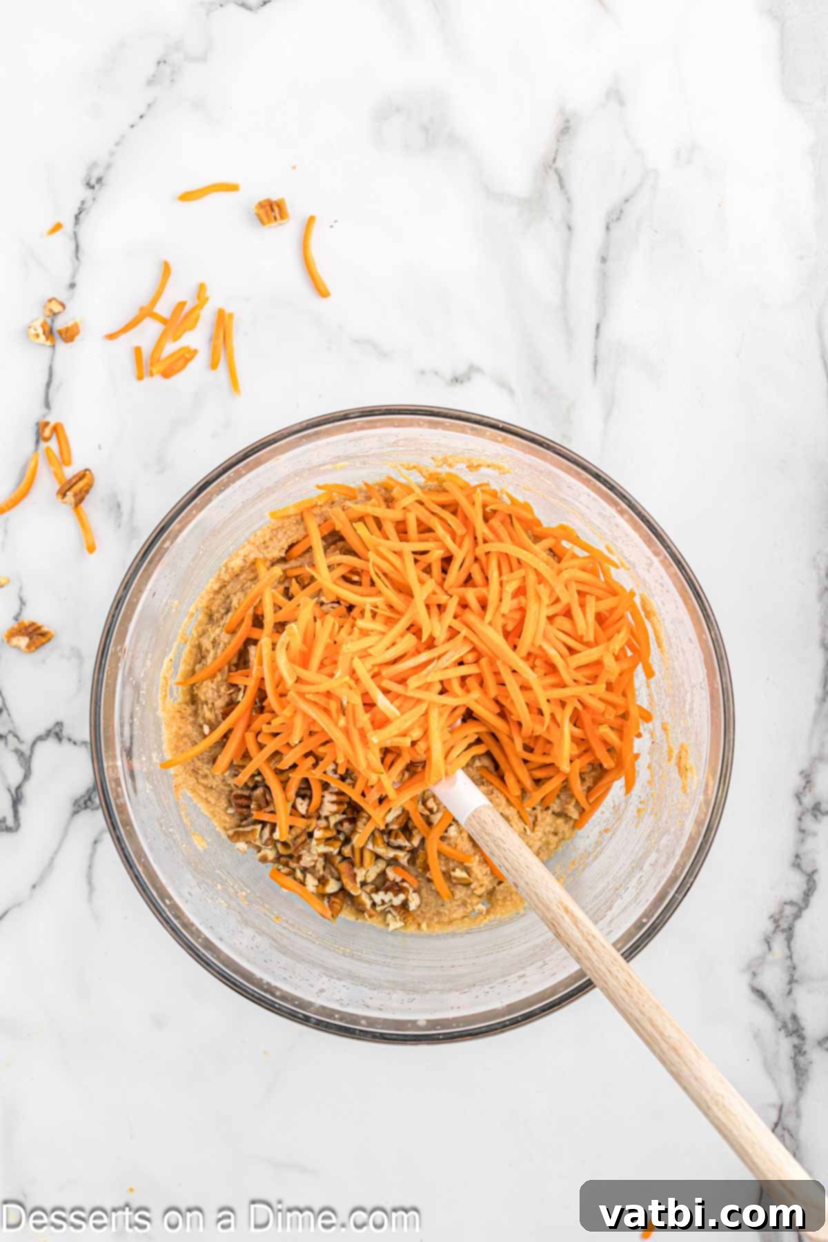 Freshly shredded carrots and chopped pecans being folded into the cake batter in a bowl, adding color and texture.