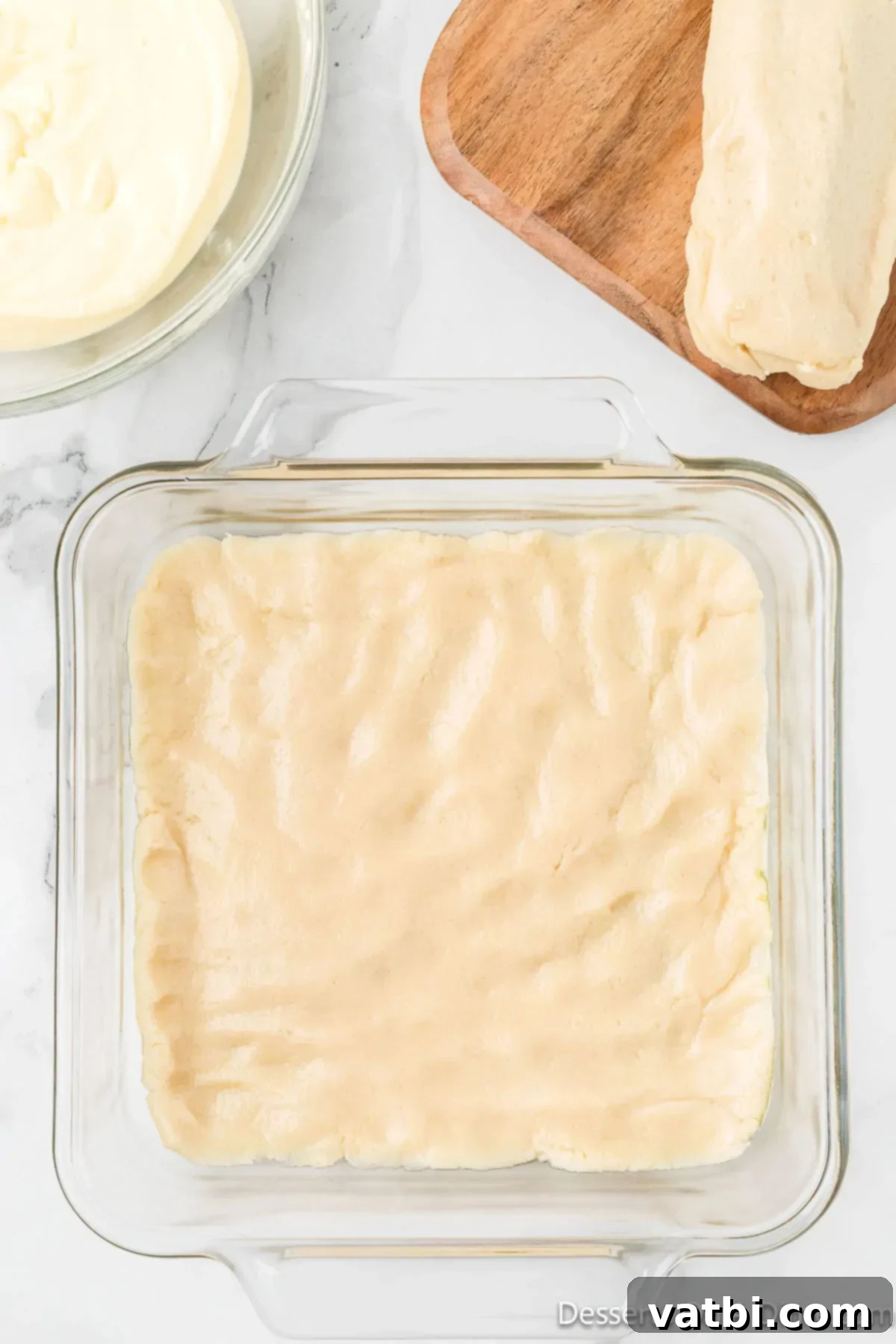 Dreamy Snickerdoodle Cheesecake Bars 6 Sugar cookie dough pressed into a baking dish. The bottom of a square baking dish is covered evenly with pressed sugar cookie dough, forming the crust.