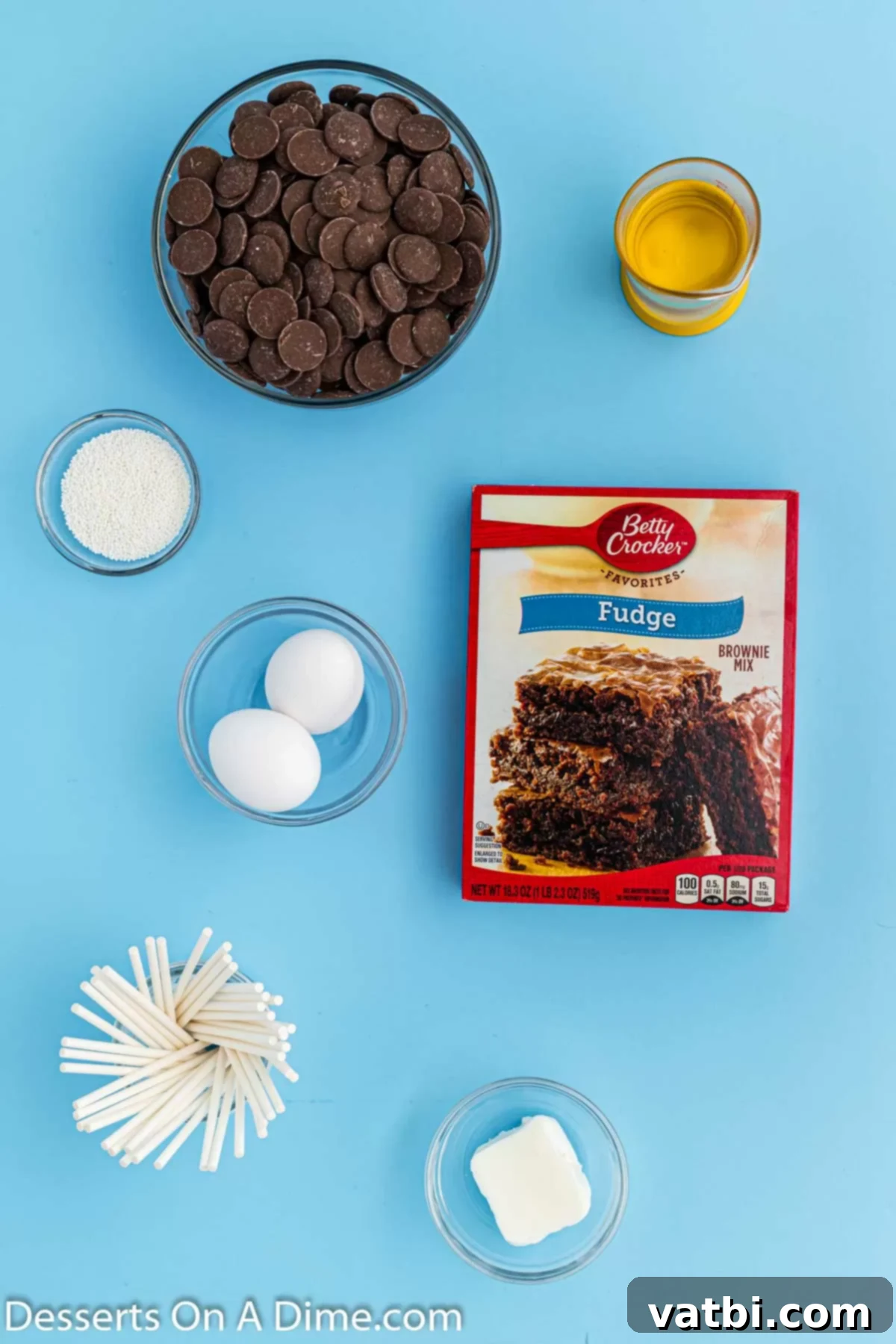 Bites of Brownie Bliss 3 A flat lay image displaying all the key ingredients for brownie cake pops: a box of brownie mix, cake pop sticks, various colors of candy melts, shortening, and white sprinkles.