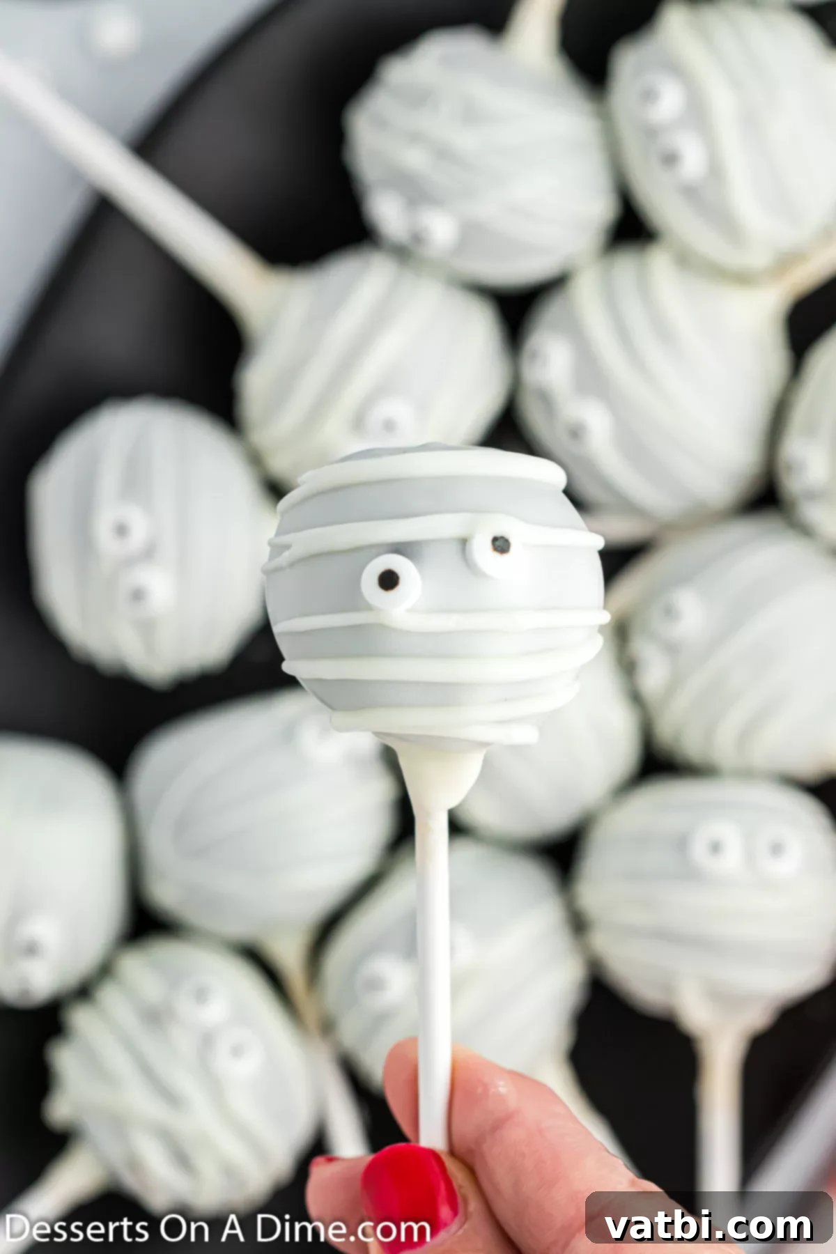 A vibrant platter of finished mummy cake pops, with one held up close to show its adorable candy eyes and chocolate wrappings.
