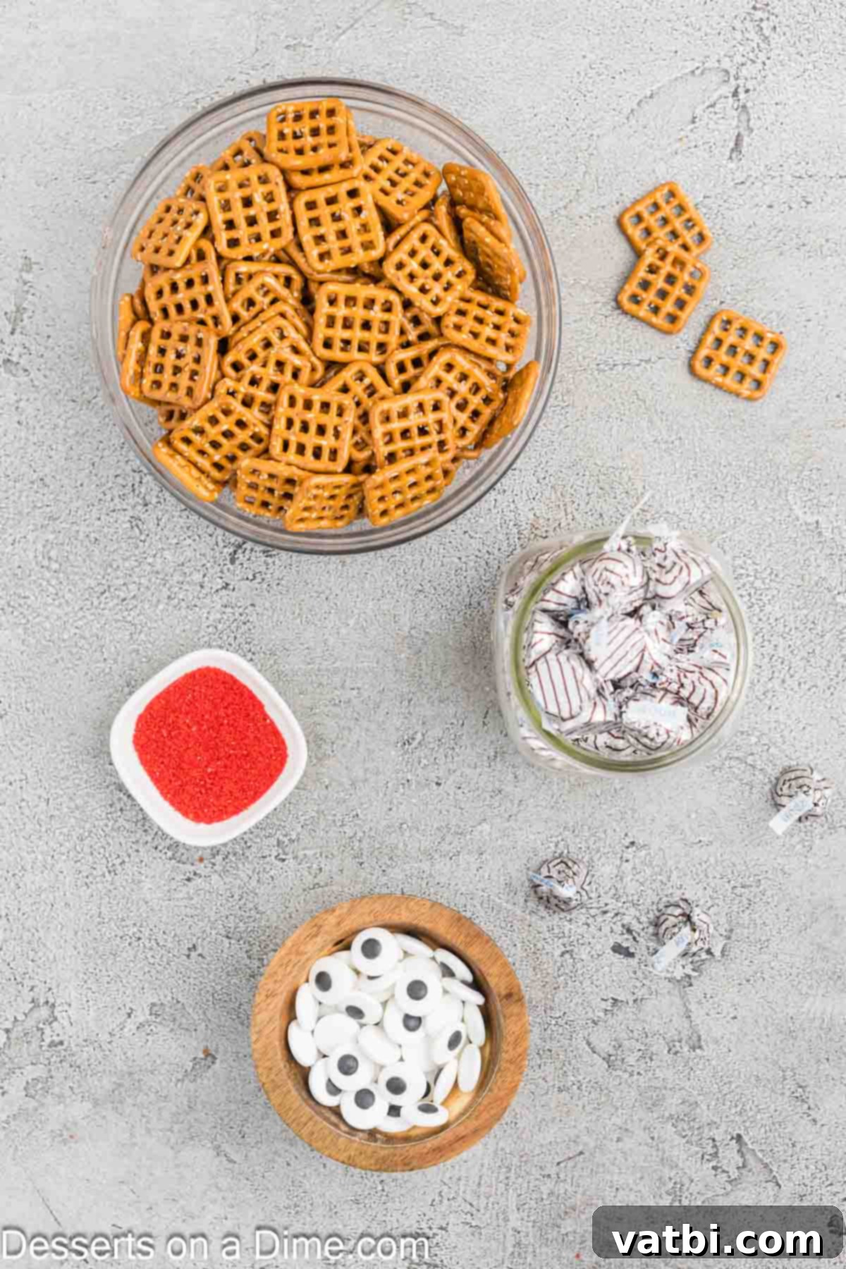A selection of ingredients for Halloween Pretzel Monster Eyeballs, including square pretzels, Hershey Hugs, candy eyeballs, and red sanding sugar, neatly arranged on a white background.