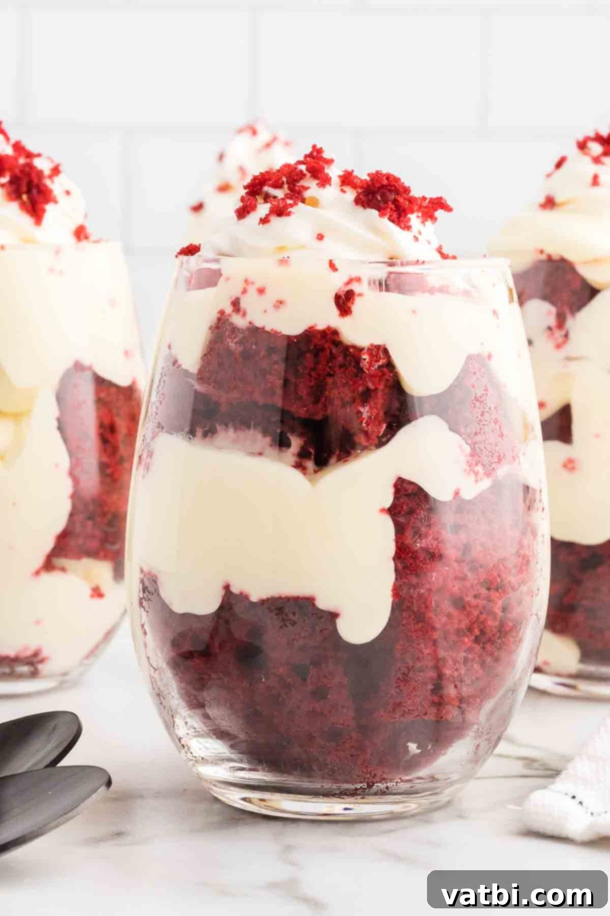Red Velvet Dream Cups 2 Close-up of a beautifully layered Red Velvet Trifle Cup, showcasing the vibrant red cake, white cream cheese filling, and fluffy whipped cream topping, garnished with cake crumbs.