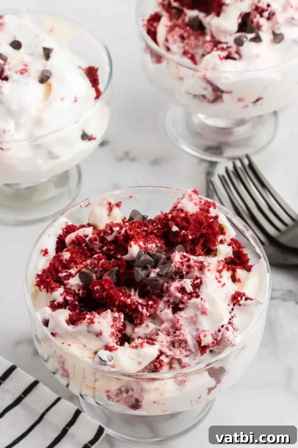 Crimson Velvet Trifle 18 Close-up of a beautifully layered Red Velvet Trifle in a clear glass serving dish, showing the distinct red cake and white cream cheese layers, garnished with chocolate chips.