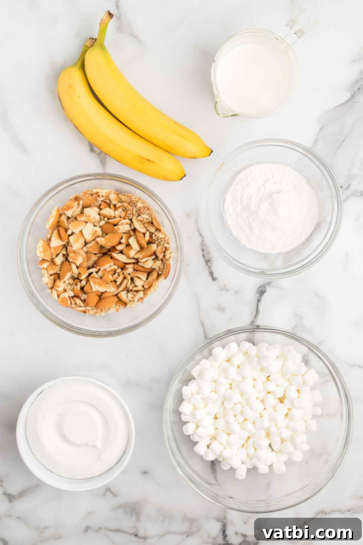 Whipped Banana Bliss 3 A flat lay photograph showcasing the key ingredients for Banana Pudding Fluff: a box of banana instant pudding mix, a carton of milk, a tub of Cool Whip, a bag of mini marshmallows, a box of vanilla wafers, and two medium-sized ripe bananas.