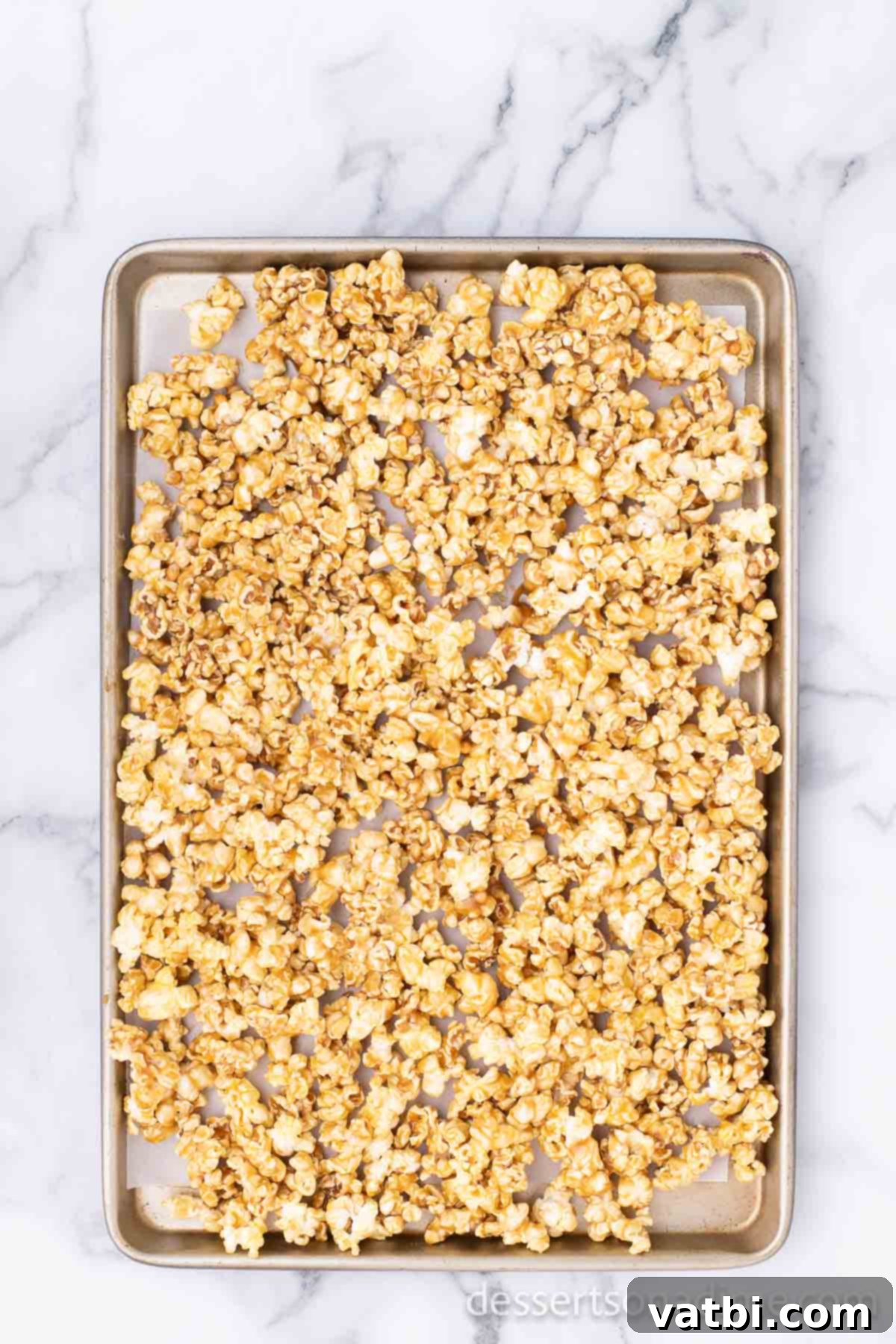 Snickers Popcorn Crunch 6 Caramel-coated popcorn spread out in a single layer on a large baking sheet, ready for baking.