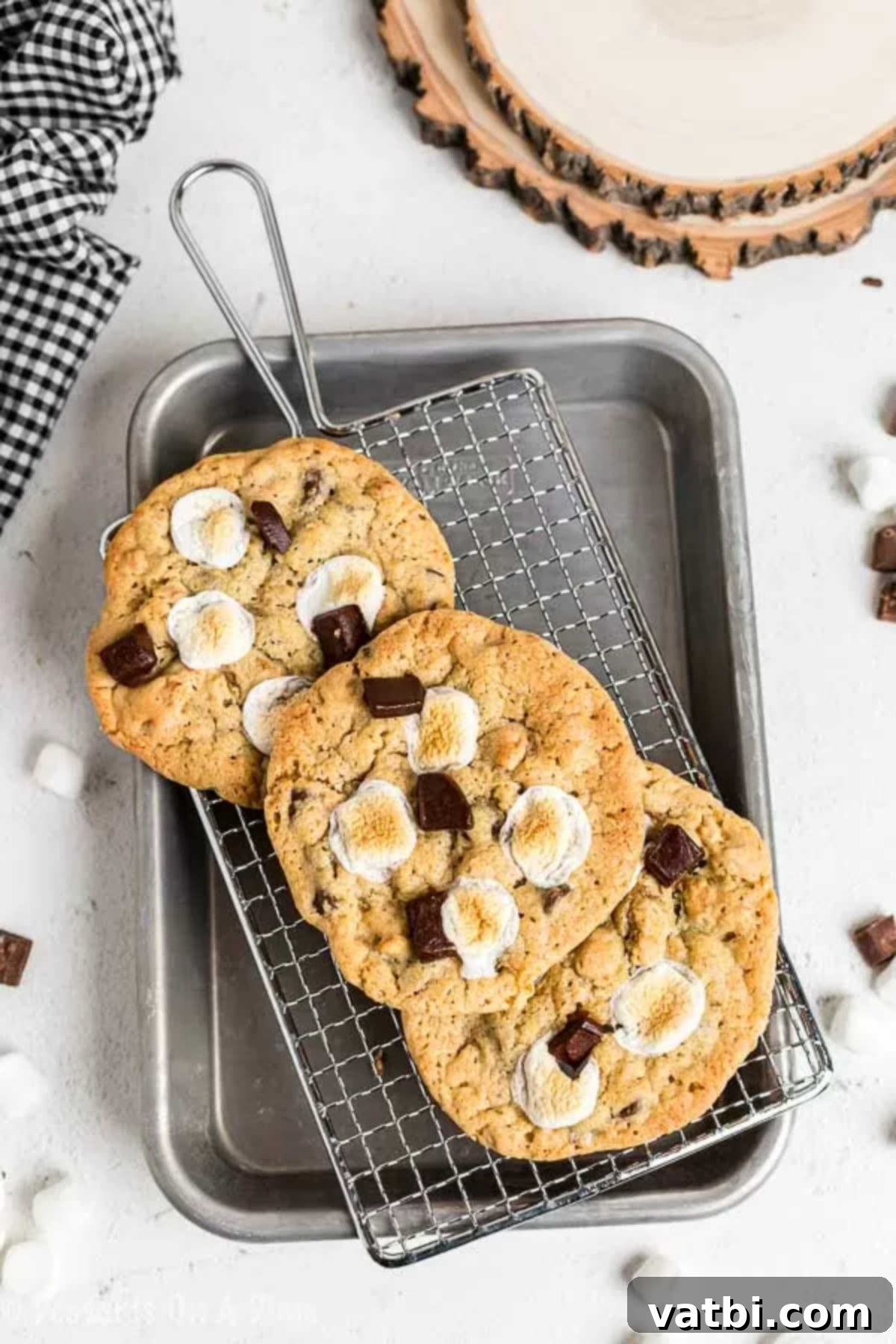 Everything you love about s'mores is packed into this tasty S'mores Cookies Recipe. Each bite is ooey gooey with lots of chocolate, marshmallows and more! Close up image of s'mores cookies sitting on a cooling rack. Golden brown cookies topped with melted marshmallows and chocolate squares.