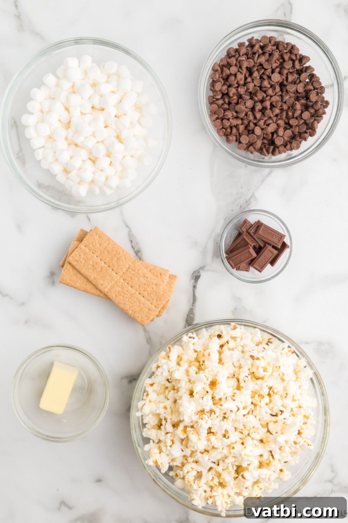 S'mores popcorn ingredients laid out: popped popcorn, a stick of butter, milk chocolate chips, mini marshmallows, graham crackers, and a Hershey's chocolate bar.