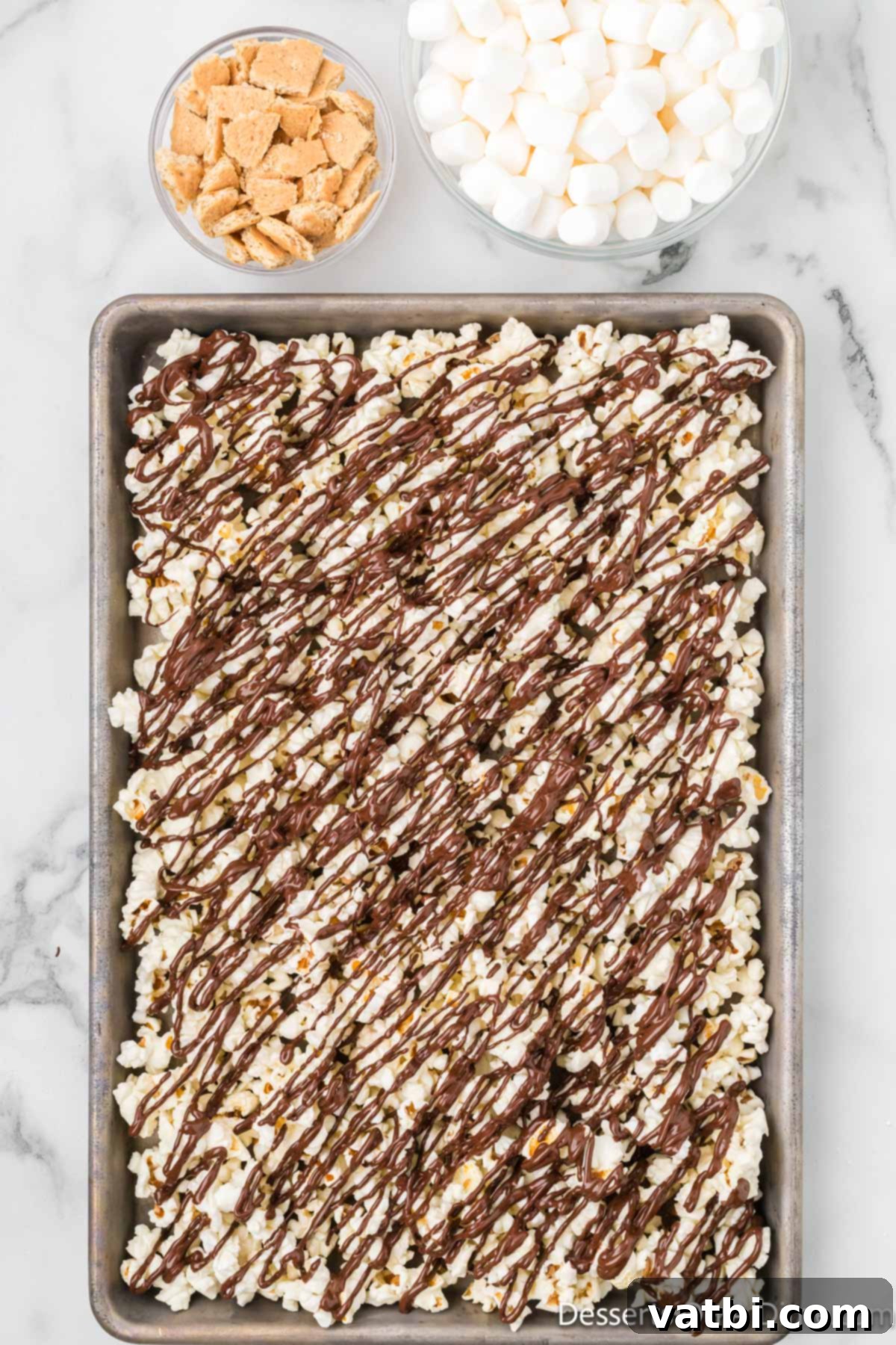 Melted chocolate generously drizzled over the spread-out popcorn on a baking sheet.