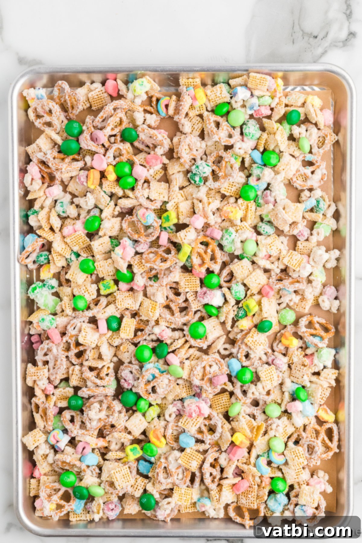 Emerald Enticement 7 White chocolate coated cereal mixture spread evenly on a baking sheet lined with parchment paper, topped with extra M&Ms and marshmallows.