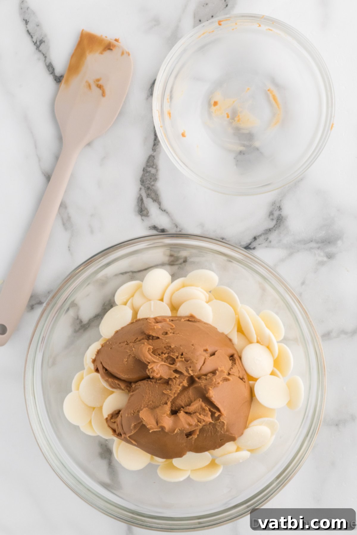 Creamy cookie butter is being added to a bowl of white melting chocolate, preparing the two main coating ingredients for heating.