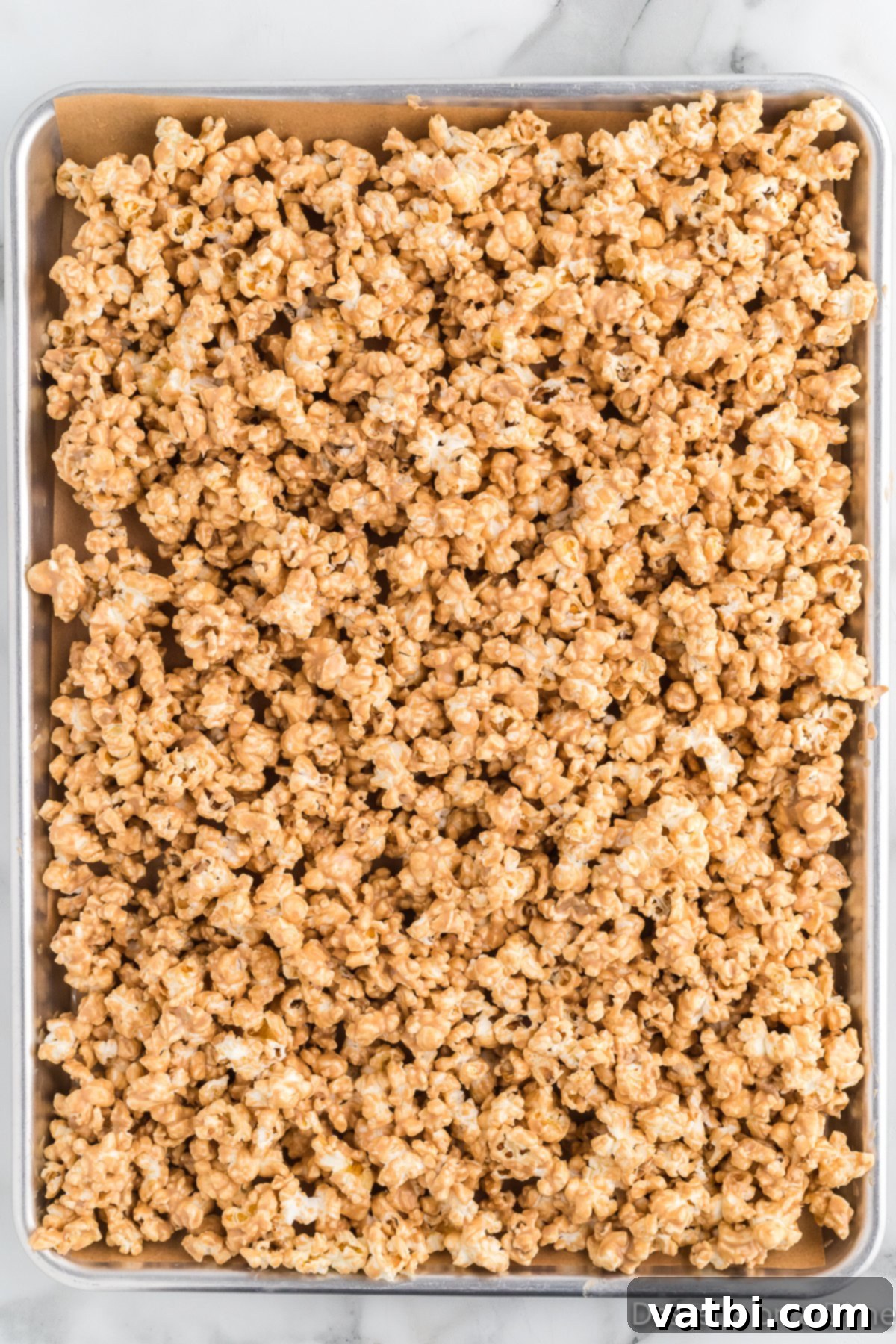 Freshly popped popcorn evenly spread on a parchment-lined baking sheet after being coated in the creamy cookie butter and white chocolate mixture, beginning to set.