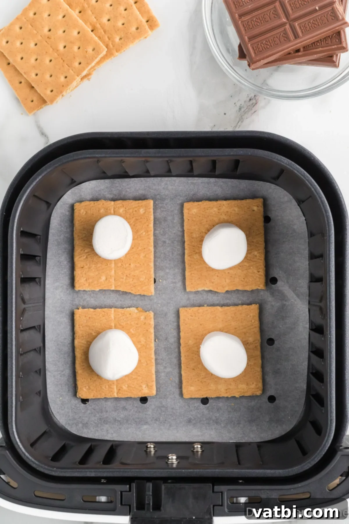 Marshmallows placed on graham cracker halves in an air fryer basket, ready for toasting.