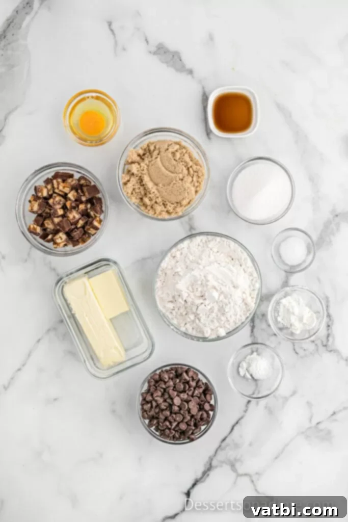 A collection of ingredients laid out on a table: butter, brown sugar, granulated sugar, all-purpose flour, baking soda, cornstarch, vanilla extract, an egg, salt, semi-sweet chocolate chips, and chopped Snickers bars.