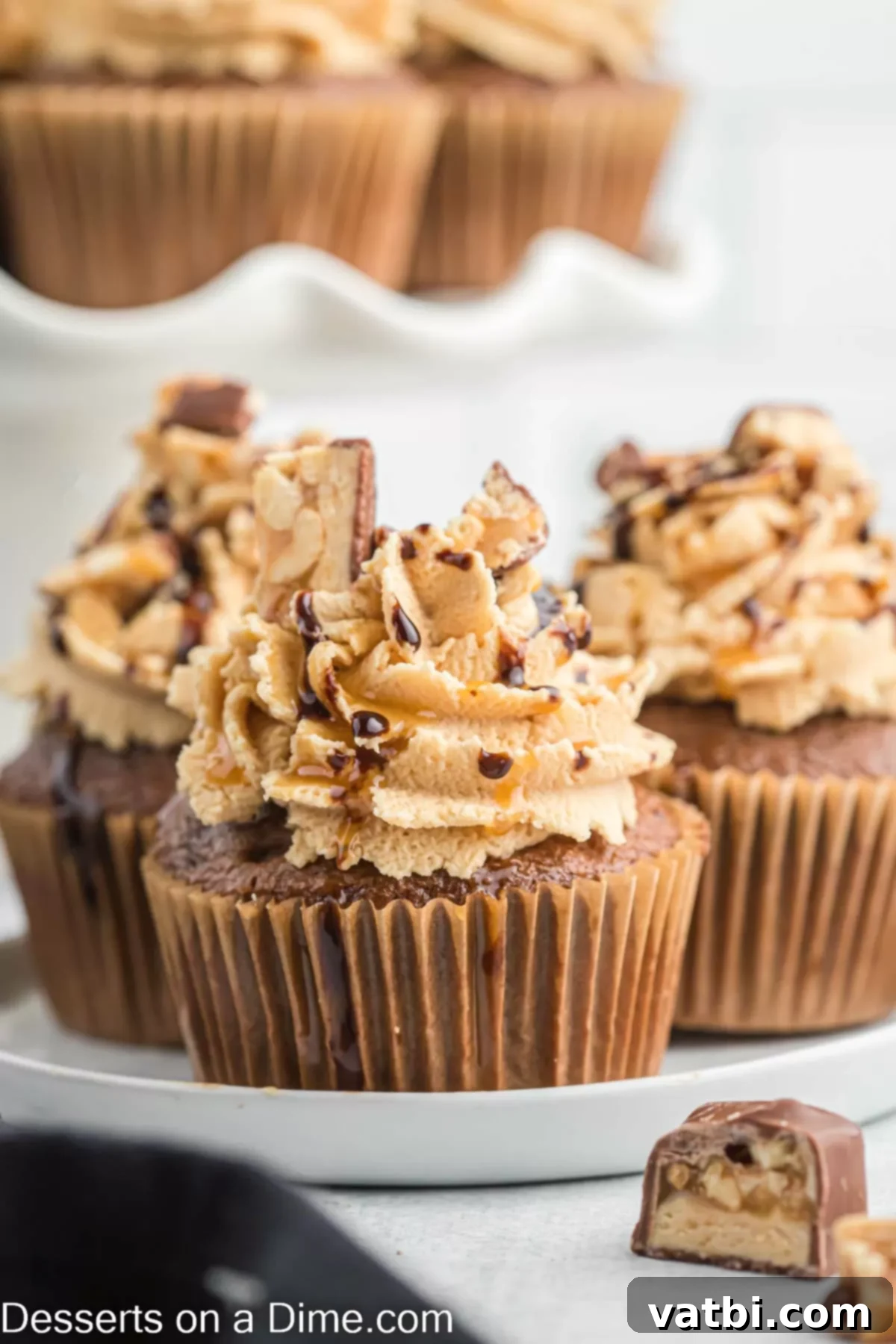 Ultimate Snickers Cupcakes 2 Snickers cupcakes on a tray, showcasing their rich chocolate base, gooey caramel-peanut filling, and fluffy peanut butter frosting, generously topped with chocolate drizzle, caramel, and mini Snickers pieces.