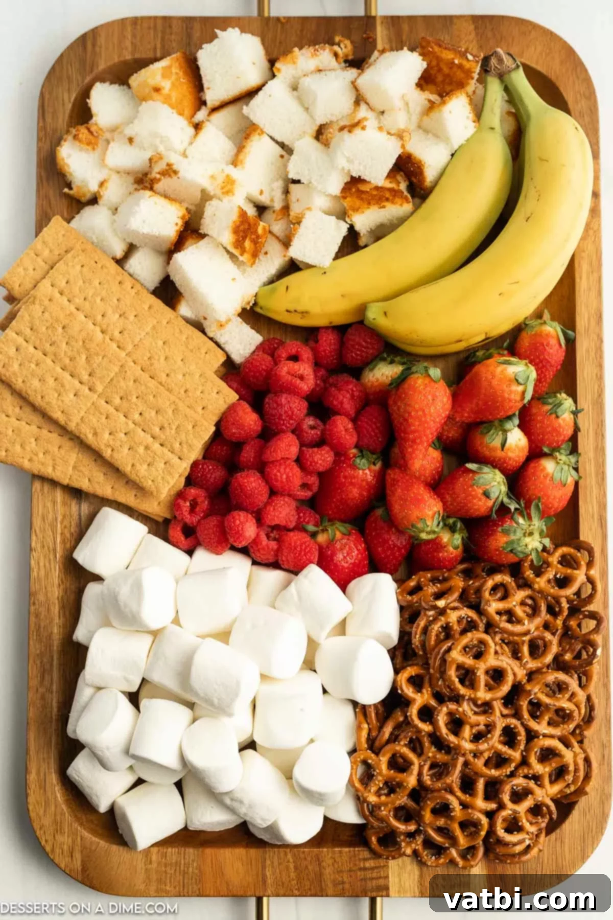 A variety of fondue dippers arranged on a board, including marshmallows, graham crackers, bananas, cake, raspberries, strawberries, and pretzels, ready for dipping into chocolate.