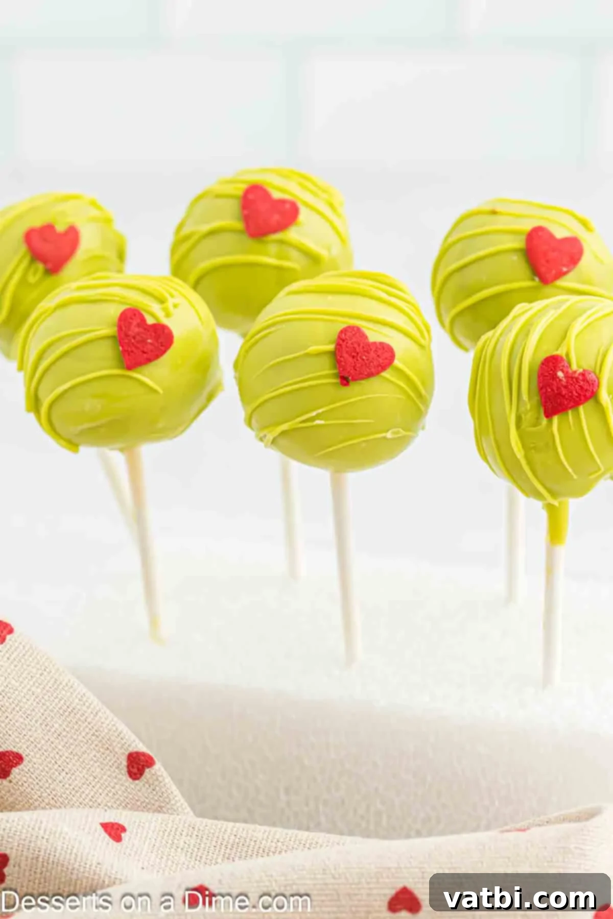 Whoville Grinch Cake Pops 2 Grinch Cake Pops on a stand, showcasing their vibrant green coating and red hearts.