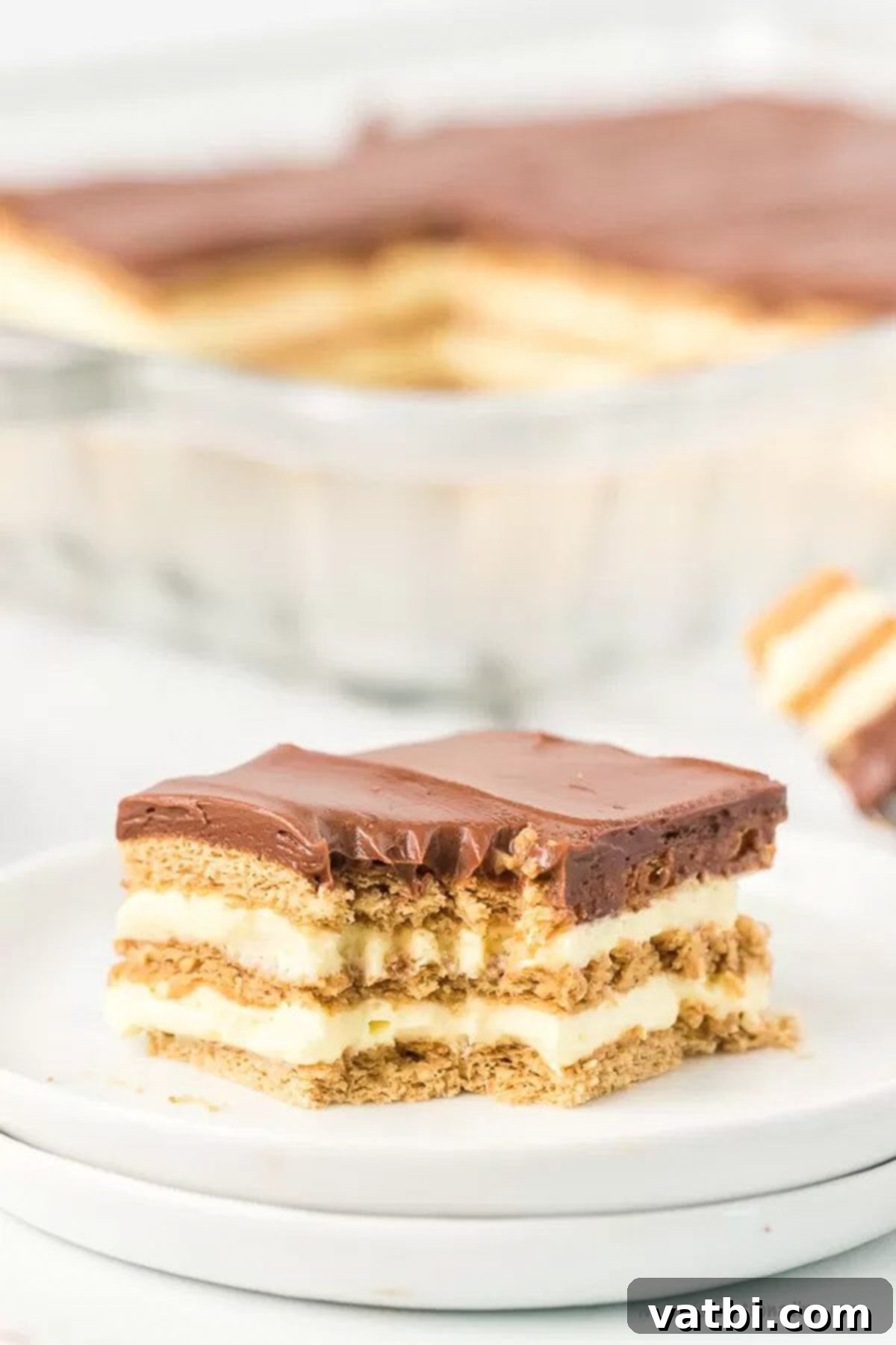 Eclair cake is an easy no bake treat with delicious layers of pudding, chocolate and graham crackers! This can be assembled in minutes for a dessert that is simple and decadent. Eclair Cake No Bake recipe is one of the best easy desserts. Anyone can make this eclair cake no bake recipe. Try eclair cake recipe today. #dessertonadime #eclaircake Close up image of slice of eclair cake on a white plate.