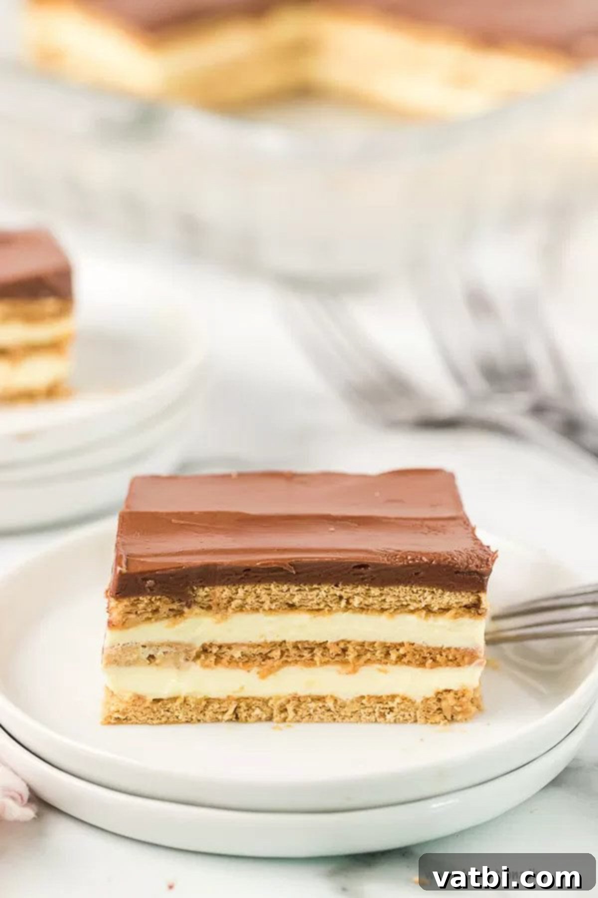 Eclair cake is an easy no bake treat with delicious layers of pudding, chocolate and graham crackers! This can be assembled in minutes for a dessert that is simple and decadent. Eclair Cake No Bake recipe is one of the best easy desserts. Anyone can make this eclair cake no bake recipe. Try eclair cake recipe today. #dessertonadime #eclaircake Close up image of slice of eclair cake on a white plate.