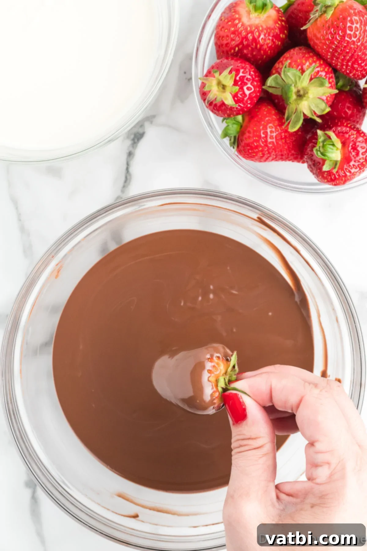 Festive Chocolate Dipped Strawberries 5 A fresh strawberry being dipped into a bowl of melted chocolate, held by its green stem.