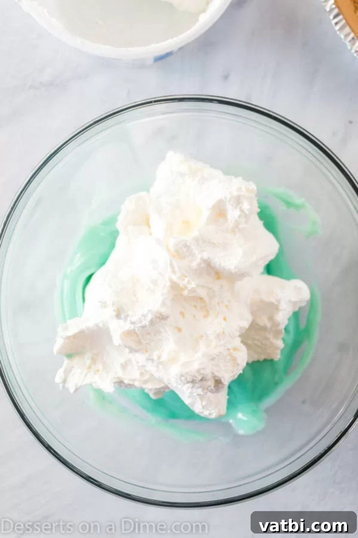 Mixing Sweetened Condensed Milk and Kool-Aid with Cool Whip A large mixing bowl containing the vibrant blue Kool-Aid pie filling mixture, with a silicone spatula gently folding in the fluffy whipped topping, creating a light and airy consistency.