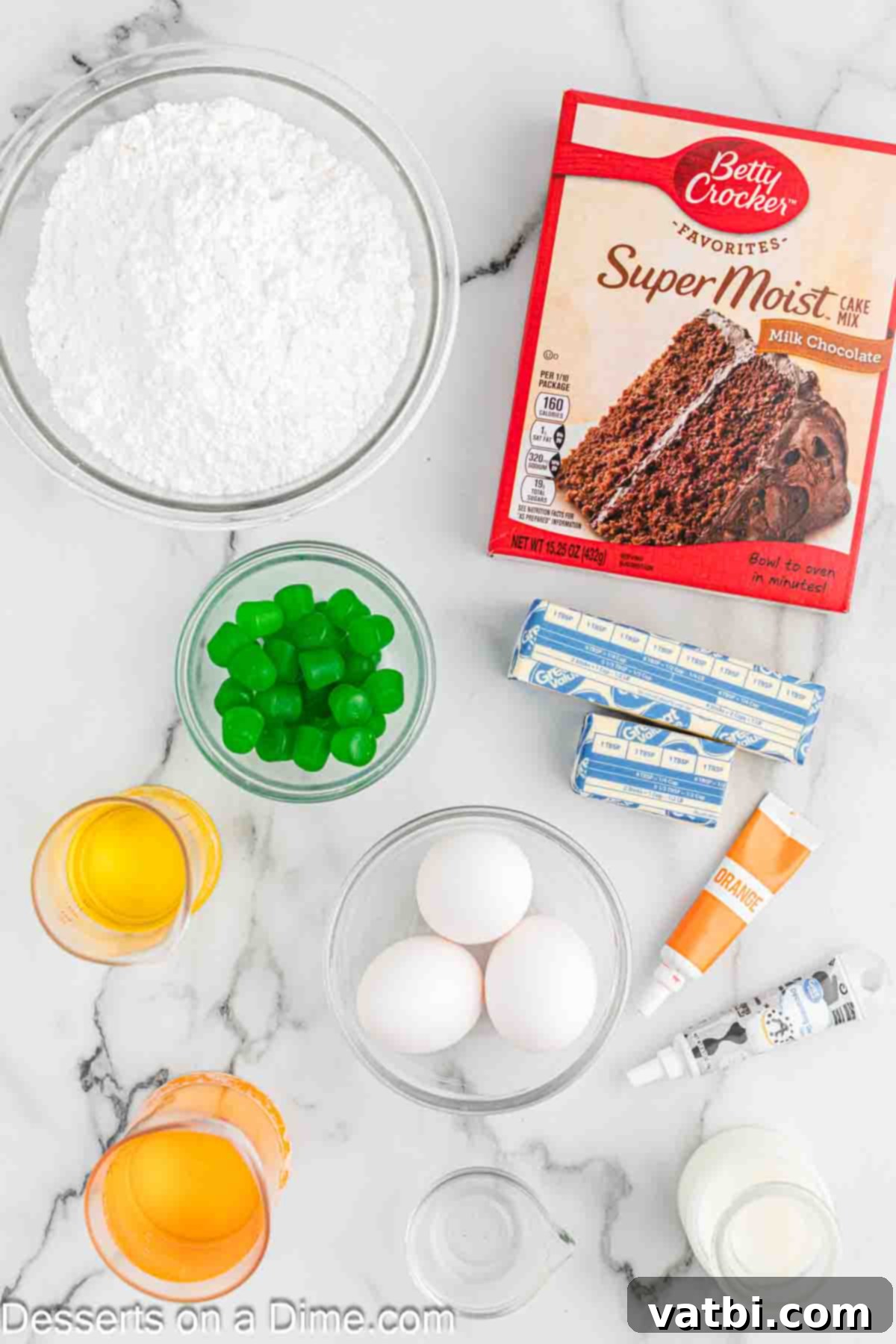 Carved Pumpkin Cupcakes 3 A flat lay image showing all the ingredients required for Jack-O-Lantern Cupcakes, including a chocolate cake mix box, powdered sugar, eggs, vegetable oil, orange gel food coloring, candy gumdrops, and butter.