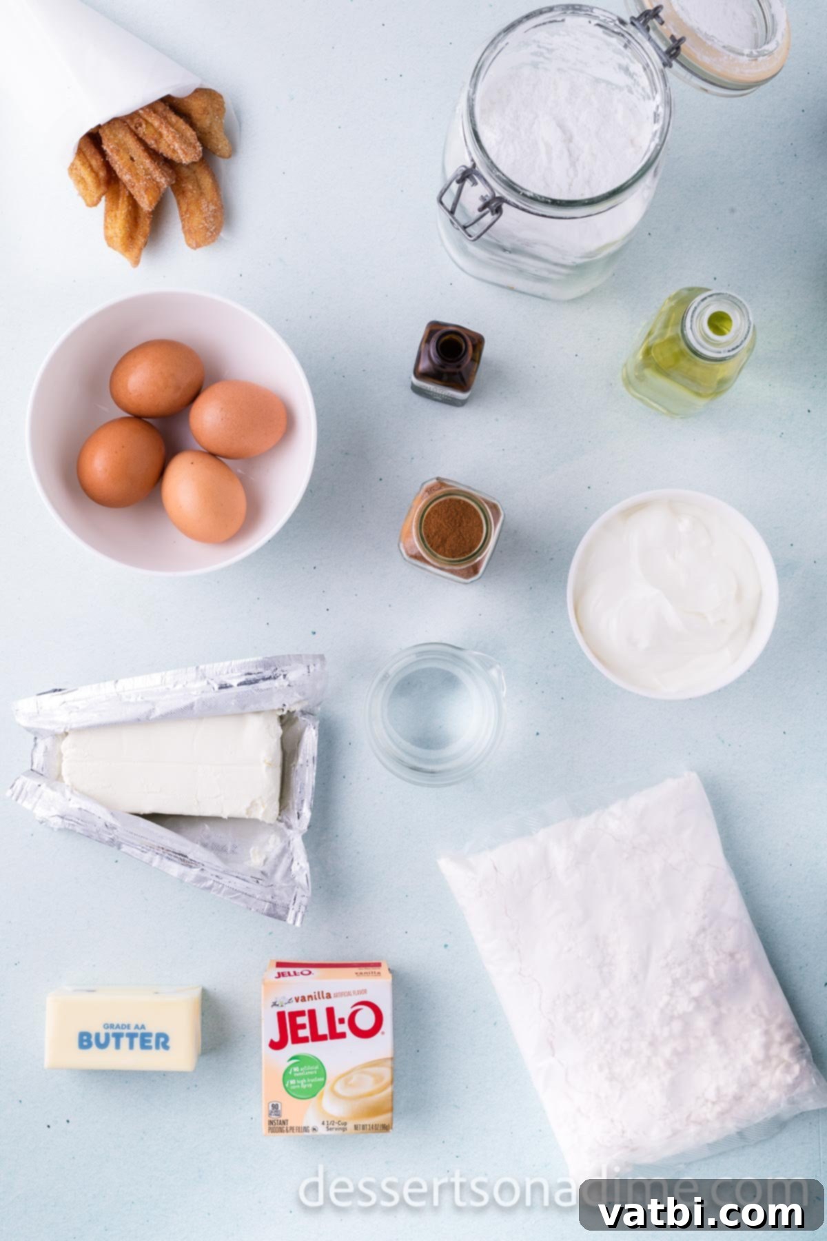 A flat lay showing all the essential ingredients for making Churro Cupcakes: a box of white cake mix, instant vanilla pudding, ground cinnamon, large eggs, sour cream, cream cheese, powdered sugar, vanilla extract, and baked churros.
