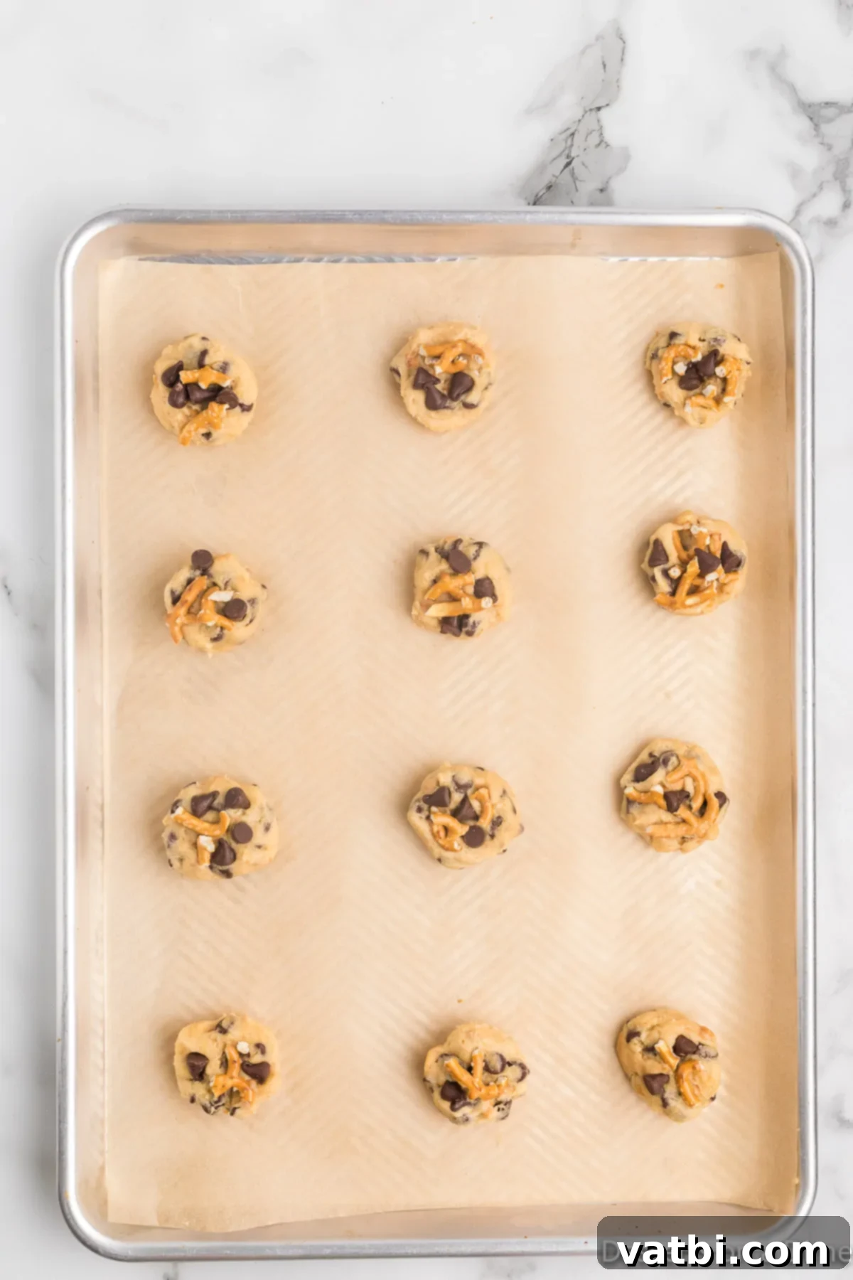 Salty Sweet Chocolate Pretzel Crunch 11 Cookie dough balls on a baking sheet, topped with additional chocolate chips and crushed pretzel pieces.