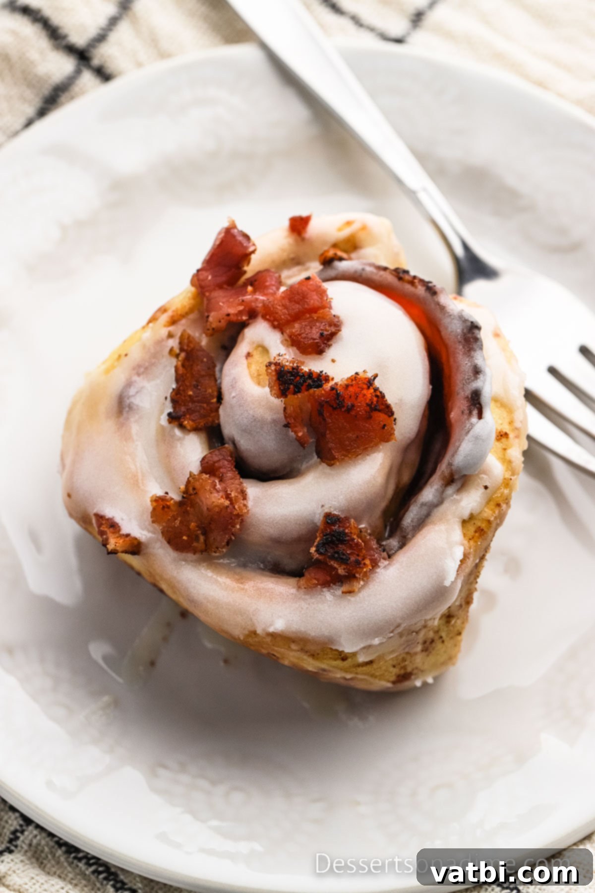 Decadent Bacon Cinnamon Buns 2 A single Bacon Cinnamon Roll on a white plate next to a silver fork, highlighting its golden-brown exterior and generous icing.
