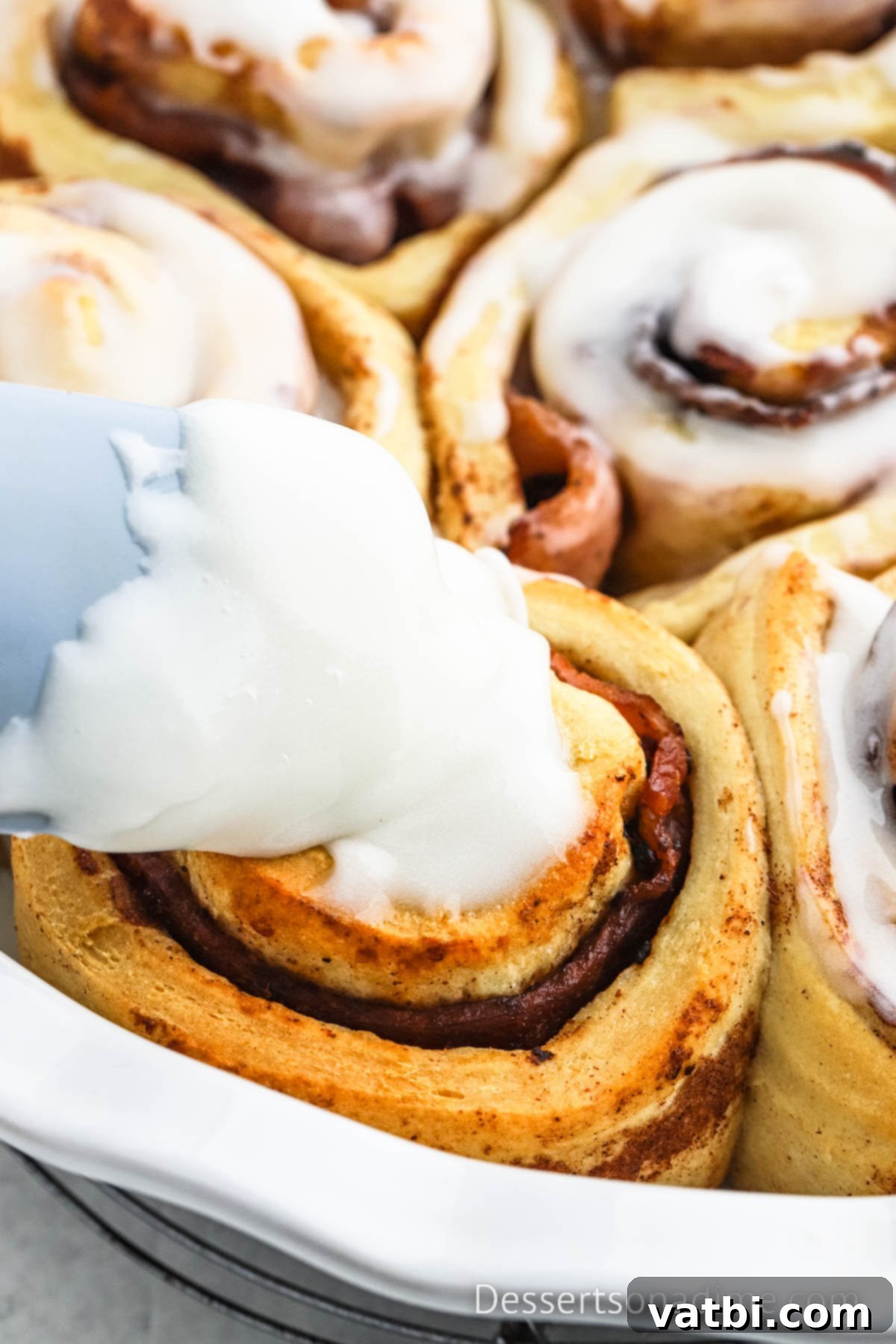 Decadent Bacon Cinnamon Buns 8 Warm, baked cinnamon rolls in a baking dish, being generously drizzled with the white cream cheese icing from their package.