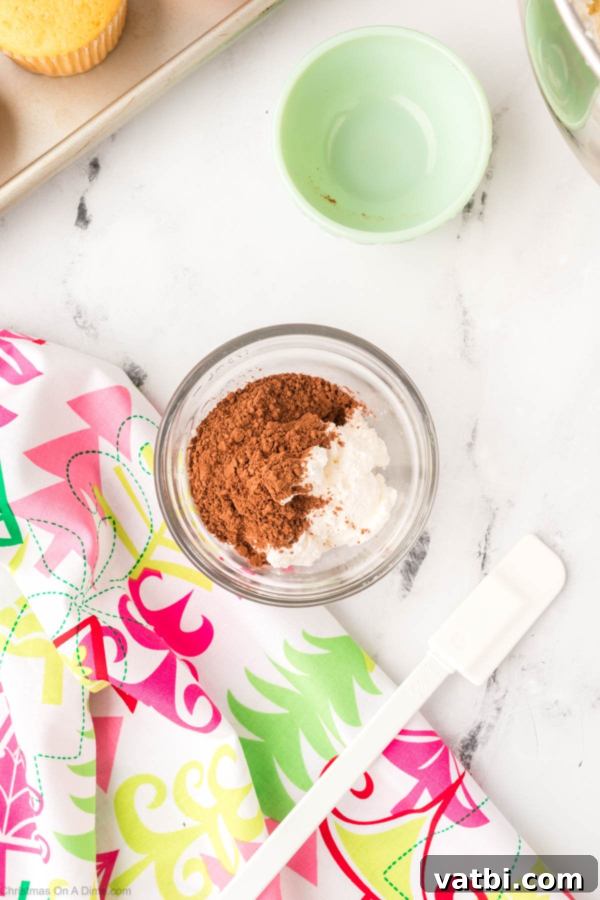 A mixing bowl with cocoa powder being added to a portion of the white frosting to create a brown color.