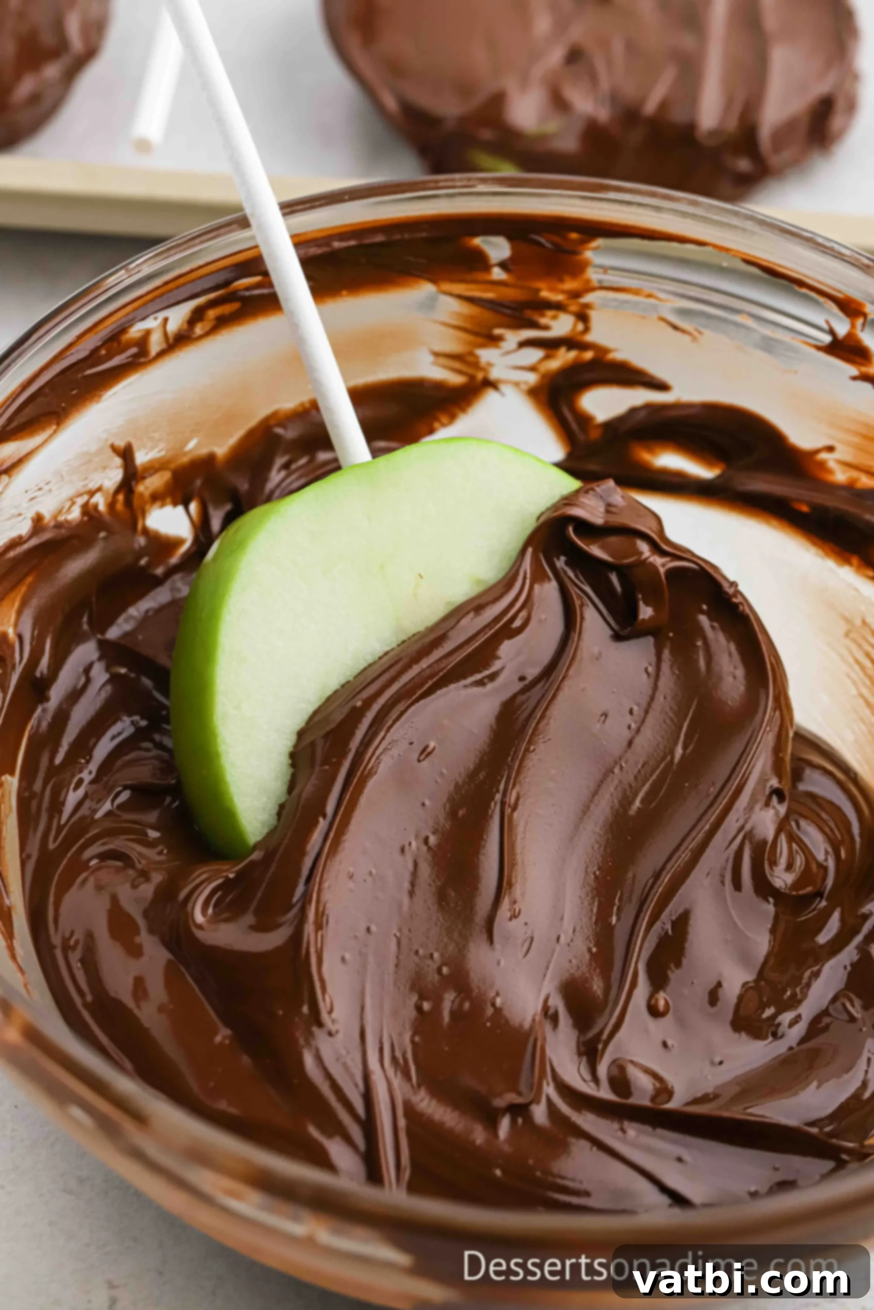 Melted chocolate in a bowl with an apple slice being coated. 