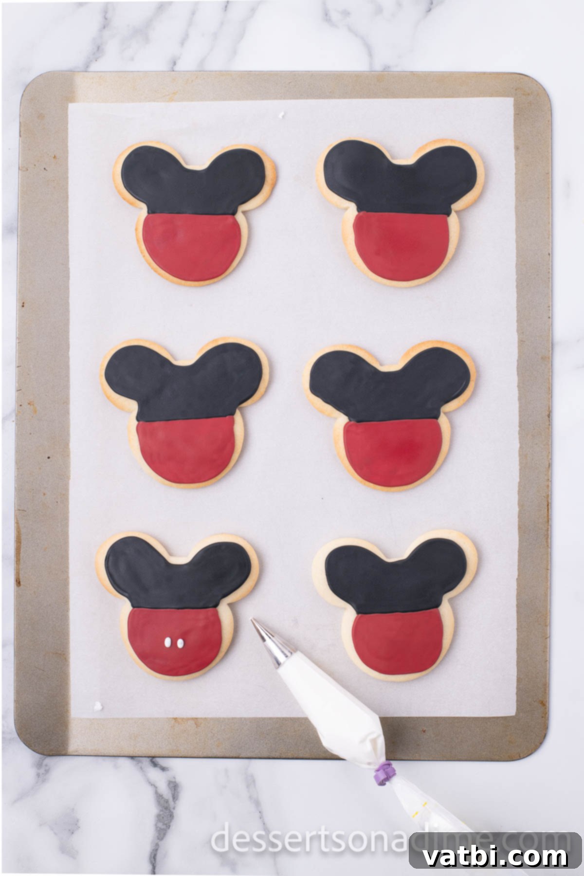 Magical Mickey Bakes 13 Mickey Mouse cookies being decorated with black and red icing using piping bags.