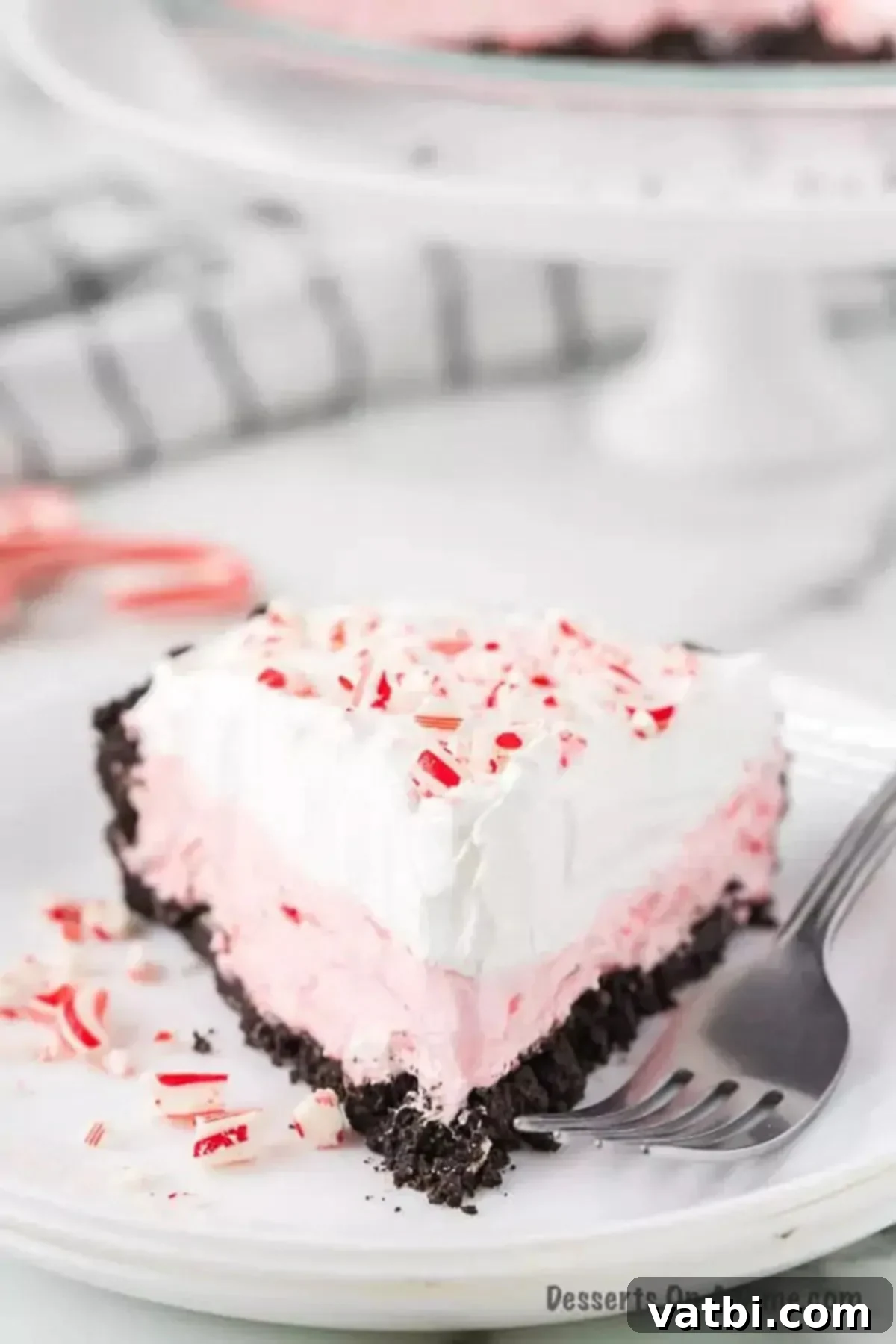 This creamy candy cane pie recipe is loaded with the best peppermint filling and oreo pie crust. Each bite of this easy no bake treat is light and fluffy. Try this tasty treat at Christmas or year round for an amazing chocolate treat. #dessertonadime #candycanepierecipe #copycat #nobake An overhead view of a slice of Candy Cane Pie on a white plate, with the remaining pie on a cake stand behind it, showcasing the festive colors and creamy texture.