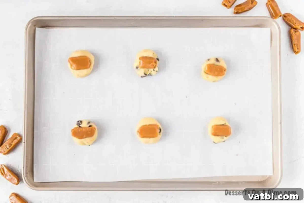 Soft caramel squares precisely placed on top of half of the portioned cookie dough balls, awaiting their top layer of dough.