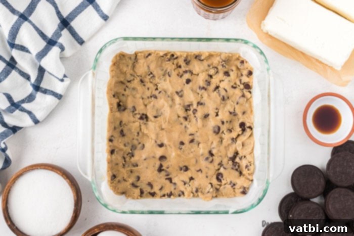 Decadent Loaded Cheesecake Bars 2 Chocolate chip cookie dough being pressed evenly into the bottom of a square baking dish lined with parchment paper.