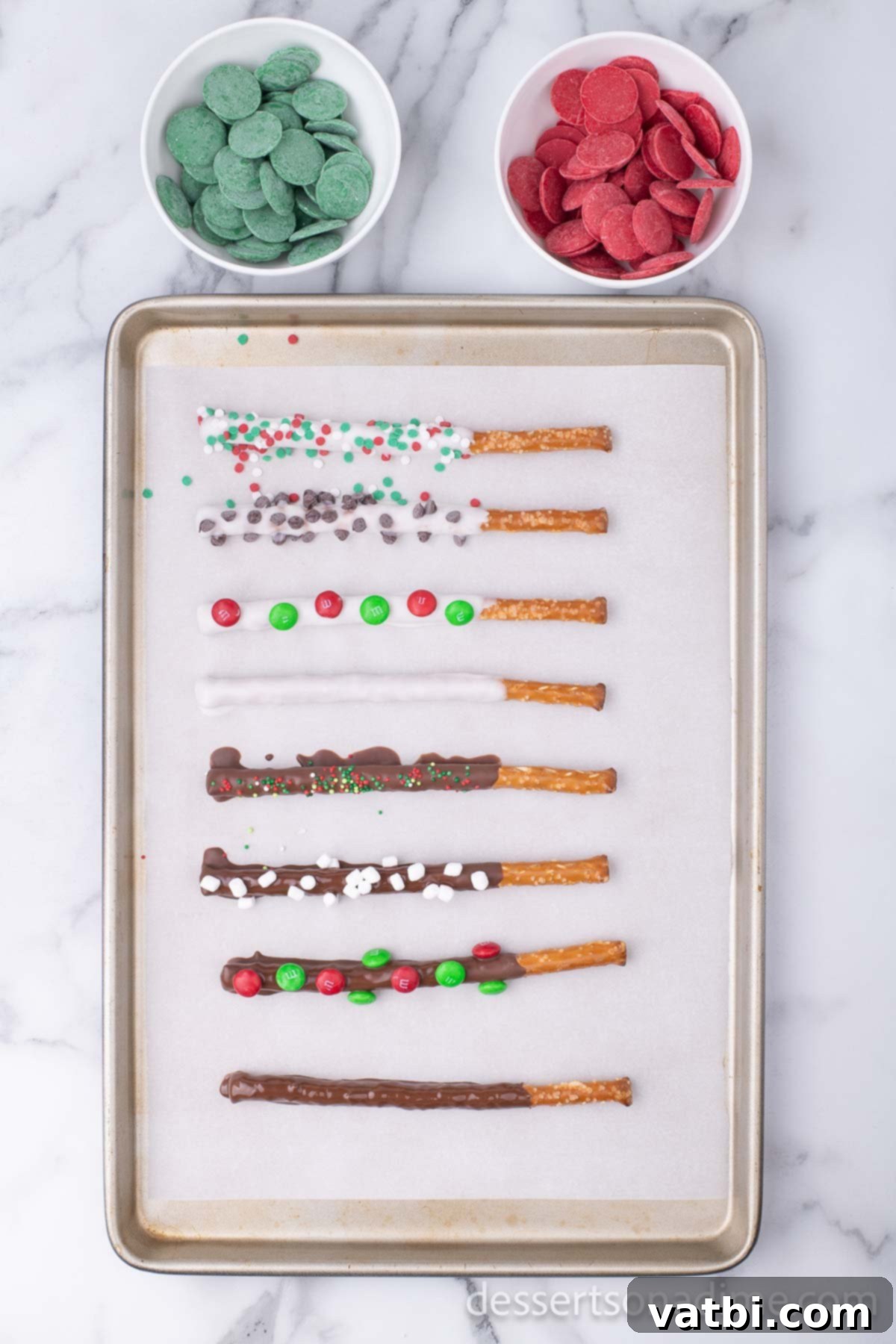 Sweet & Salty Holiday Pretzel Treats 4 Pretzel rods decorated with festive Christmas sprinkles, M&Ms, and drizzles of colored candy melts on a baking sheet