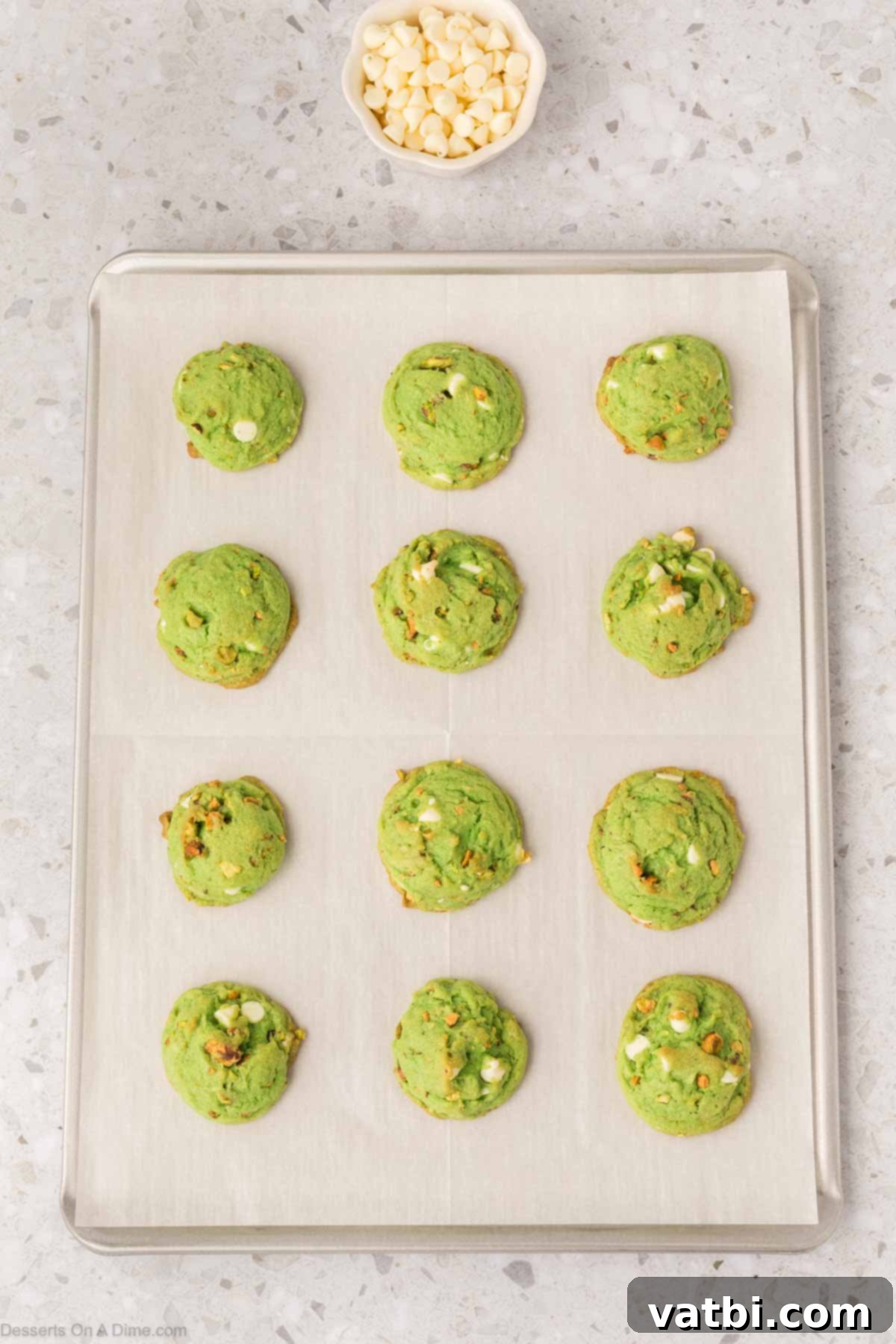 Emerald Kiss Cookies 11 Warm pistachio cookies on a baking sheet, with additional white chocolate chips pressed on top.
